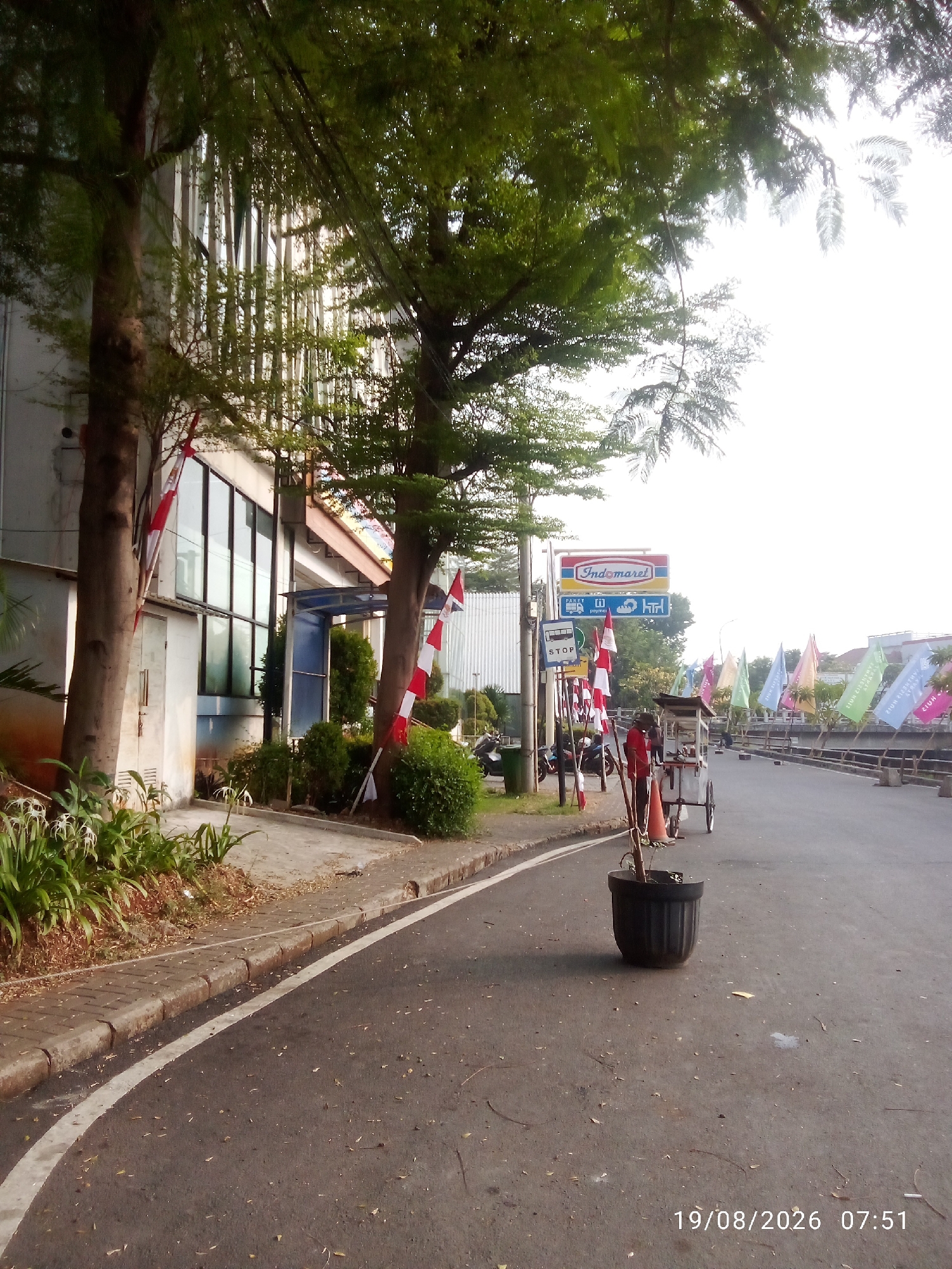 The image depicts a street scene in East Jakarta, Indonesia. On the left side of the road, there is a modern building with large glass windows, flanked by trees with red and white flags tied to their trunks. A sidewalk with a white line marking its edge runs along the building. Further down the street, a sign for "Indomaret" is visible, indicating a convenience store. A stop sign is also present. In the foreground, a dark pot with a plant stands on the asphalt. A street vendor with a cart is also present on the right side of the road. Numerous colorful flags are displayed along the opposite side of the street, which appears to be a pedestrian walkway or elevated area. The road is paved with asphalt and scattered with fallen leaves. The sky is bright and overcast, suggesting daytime. The timestamp "19/08/2026 07:51" is visible in the lower right corner of the image.