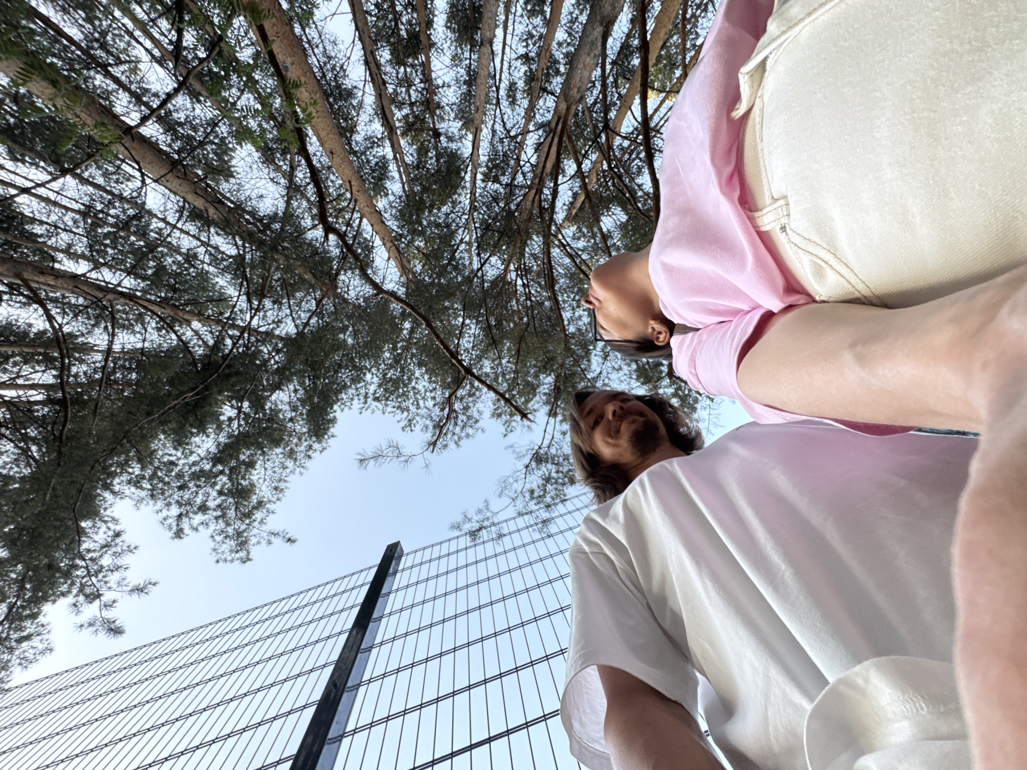 The image depicts a low-angle shot looking upwards towards two individuals standing near a wire fence and beneath trees, possibly in Izhevsk, Russia. The sky is visible through the branches. One individual is wearing a pink long-sleeved shirt and light-colored overalls. The other person is wearing a white t-shirt. The focus of the shot is primarily on the sky, trees, fence, and clothing.