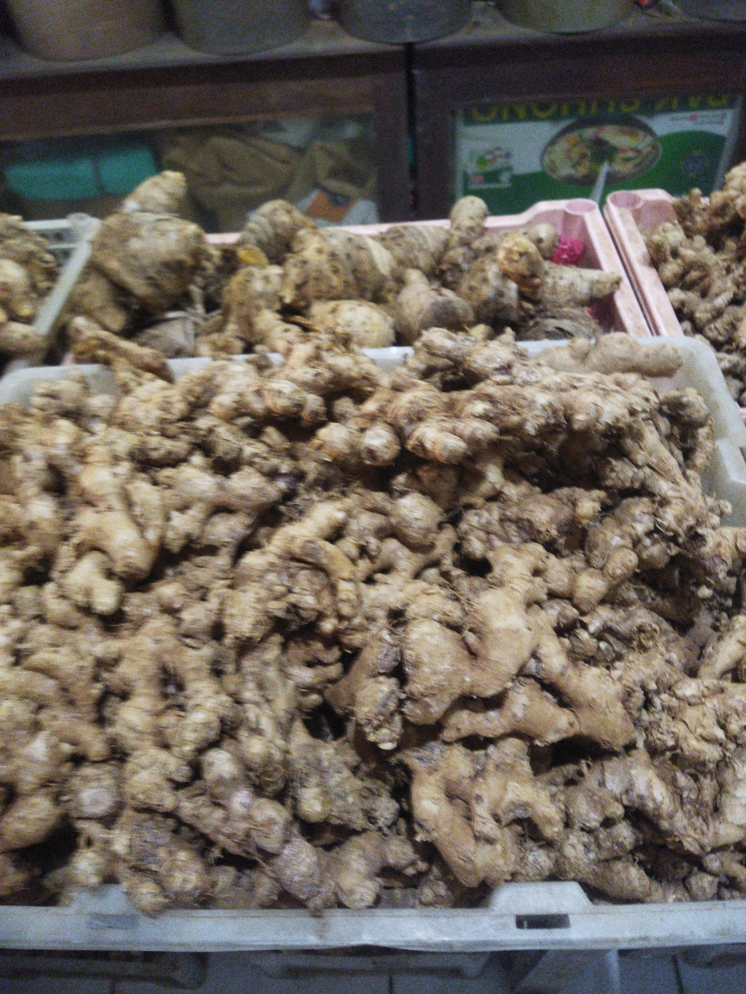 The image shows a close-up view of several crates filled with ginger. The ginger roots are light brown and appear to be whole and unpeeled. They are piled high in the crates, with some spilling over the edges. In the background, there are shelves with what appear to be other goods, possibly spices or produce. A portion of a sign with green and white text is visible in the upper right background, suggesting a market or store setting. The lighting is dim and the image quality is grainy, making it difficult to discern fine details. The scene is likely a market stall in Sukoharjo, Indonesia, given the context provided.