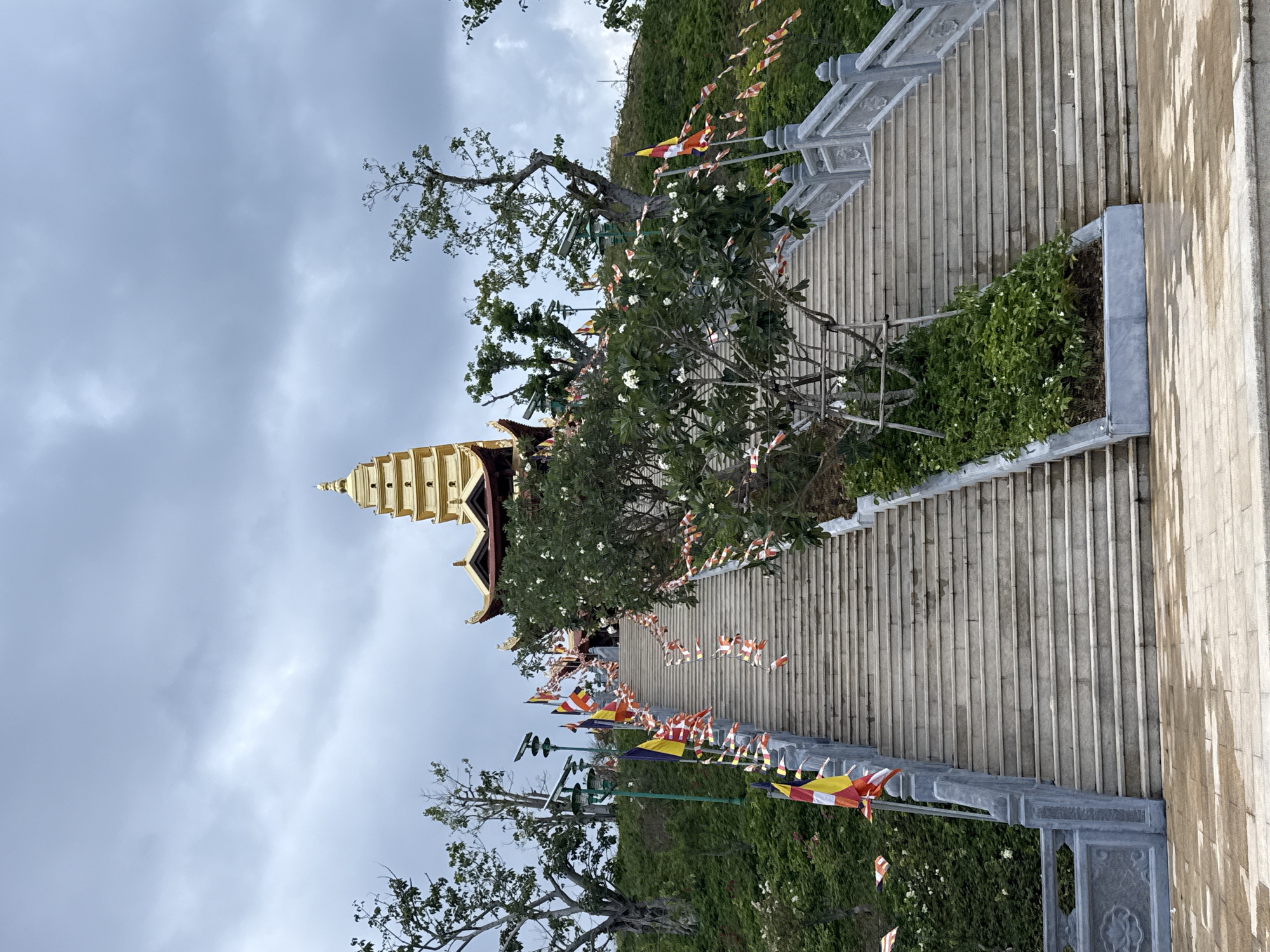 A low-angle, vertically oriented shot depicts a long, wide set of stone stairs leading up to a golden pagoda in Nam Nha Trang Ward, Vietnam. The sky is overcast with grey clouds. The stone staircase is lined with green vegetation and trees, interspersed with small, colorful prayer flags. The pagoda is visible in the upper left quadrant of the image, its golden tiers rising toward the sky. To the right of the stairs is a stone pathway with a slight sheen, suggesting recent rain or washing.