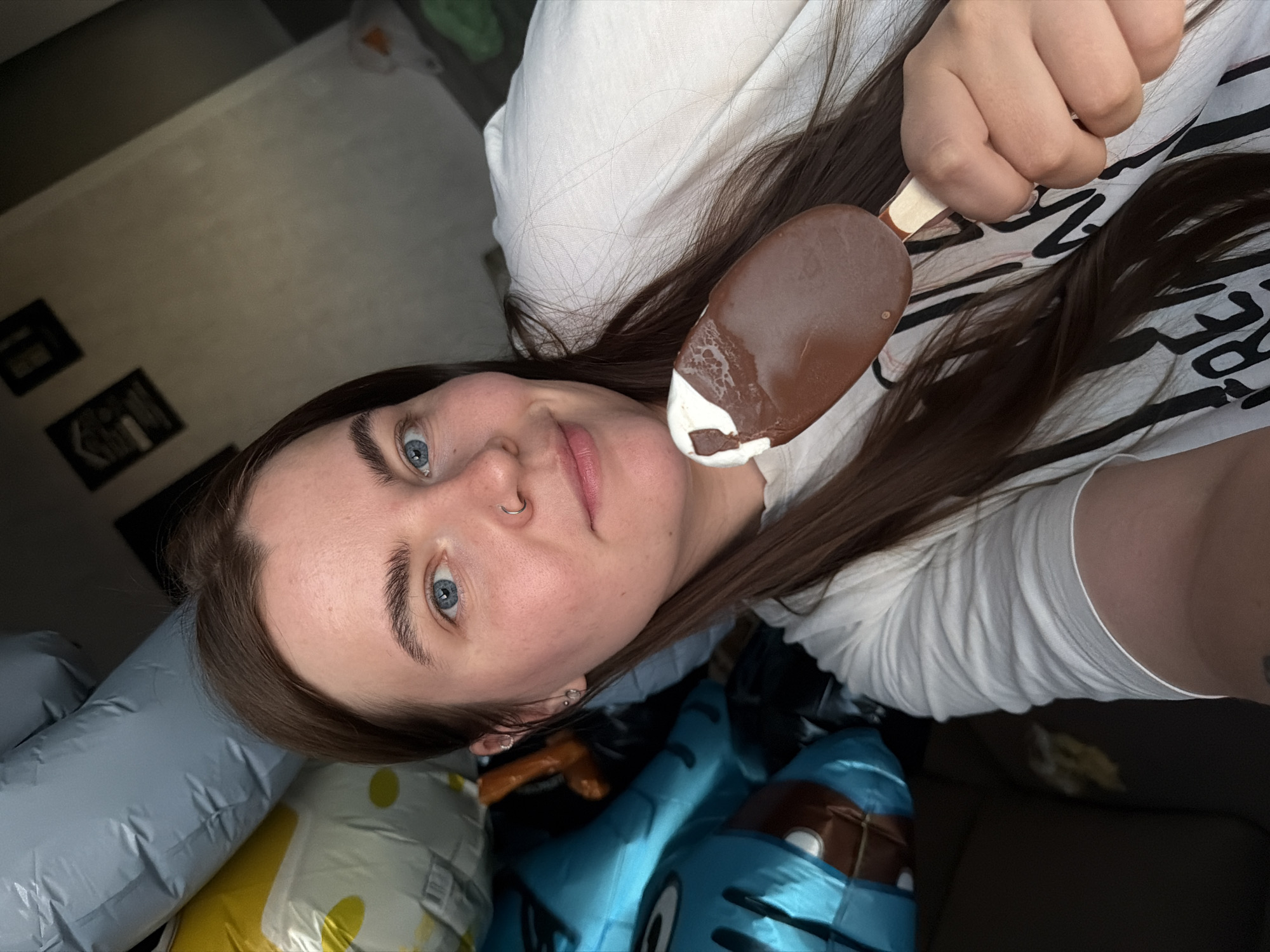 In the city of Chaikovsky, Russia, a young woman with long, brown hair and blue eyes is captured in a selfie. She is wearing a white t-shirt and holding a chocolate-covered ice cream bar with a bite taken out of it. Her expression is a gentle smile, and she has a nose ring and earrings. Behind her, partially visible, are several deflated balloons, one blue and cartoon-like, another yellow, and a large gray one. The background suggests an indoor setting, possibly a living room, with framed pictures on the wall and a hint of a couch. The lighting indicates it may be daytime, with sunlight streaming in, creating shadows.