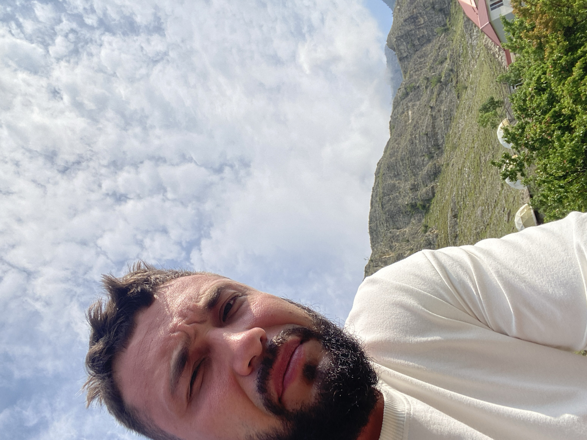 A close-up, high-angle selfie shows a man with dark hair and a beard. He is wearing a white t-shirt. The background features a bright, cloudy sky, a rocky, green mountain slope, and some trees. In the lower right corner, a pink-roofed building and several geodesic domes are visible. The scene is consistent with an outdoor location in Chalda, Russia.