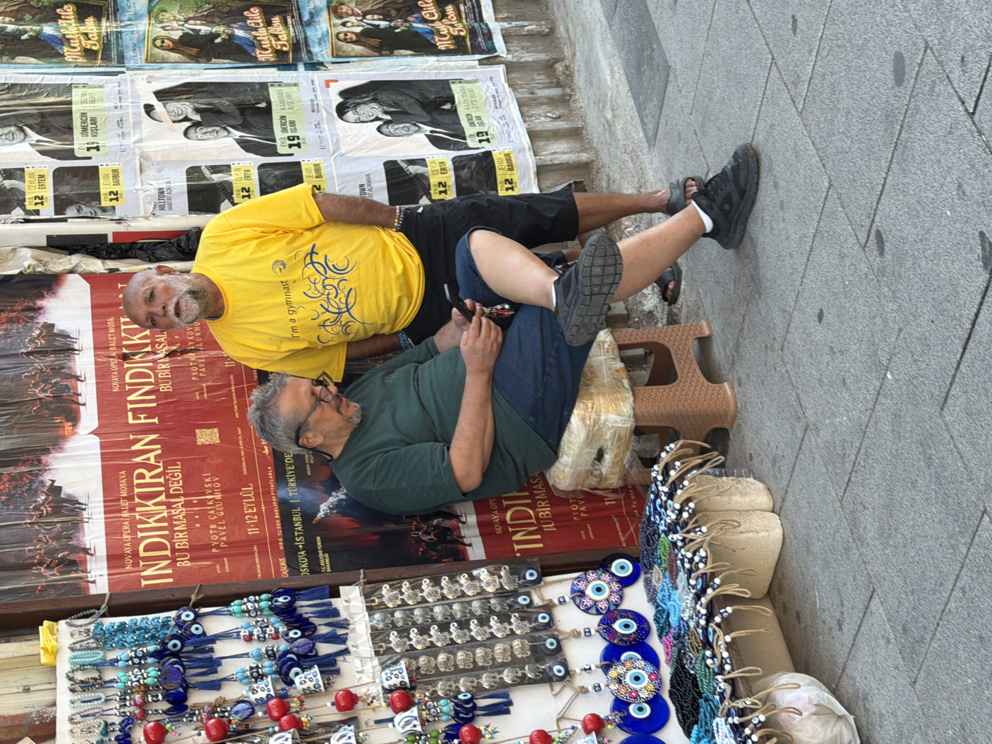 Two men are seen on a sidewalk in Kadıköy, Turkey, with posters displayed behind them. The man on the left, wearing glasses and a dark green t-shirt, sits on a stool with a blue cellphone in his hand. He is wearing blue jeans and black sneakers. The man on the right, wearing a bright yellow t-shirt with the text "I'm a gymnast" and black shorts, stands beside him. He is barefoot. A display of beaded bracelets and evil eye charms is visible in the foreground to the left. The posters behind the men appear to be advertisements, one with images of performers in red and black. The ground is paved with gray stone tiles.