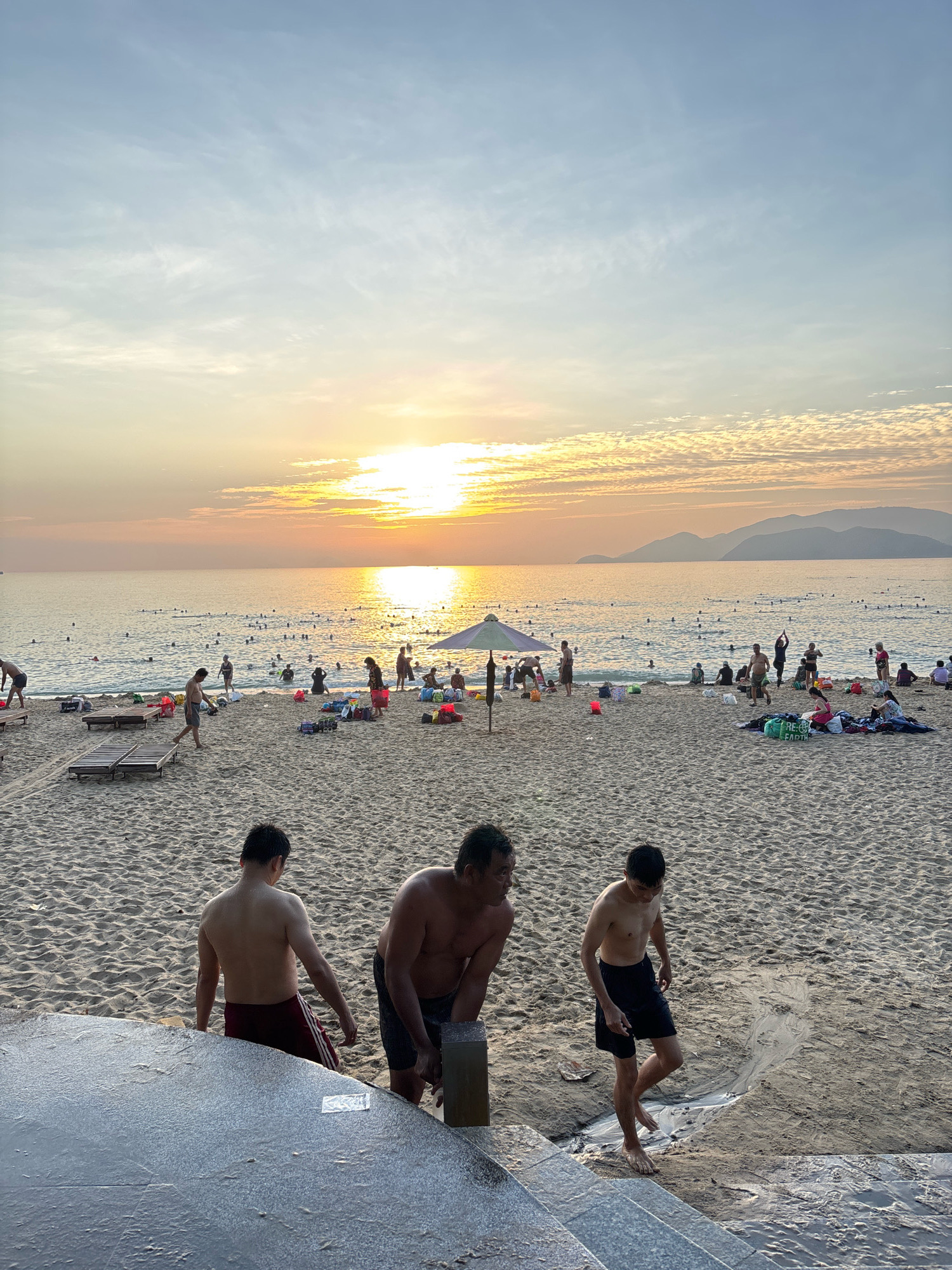 The image depicts a serene sunset scene on a crowded beach in Nha Trang, Vietnam. The sky is awash with hues of orange and yellow as the sun dips below the horizon, casting a warm glow over the ocean and the sand. A multitude of people are scattered across the beach and in the water, enjoying the last rays of daylight. Some are swimming, others are lounging on beach chairs or towels, and children are playing in the sand. The foreground is dominated by a concrete structure, possibly a set of stairs or a pathway, with three individuals in swimwear walking away from the viewer. In the background, the calm sea stretches to meet a silhouetted mountain range. The overall atmosphere is one of relaxation and leisure, characteristic of a popular tourist destination.