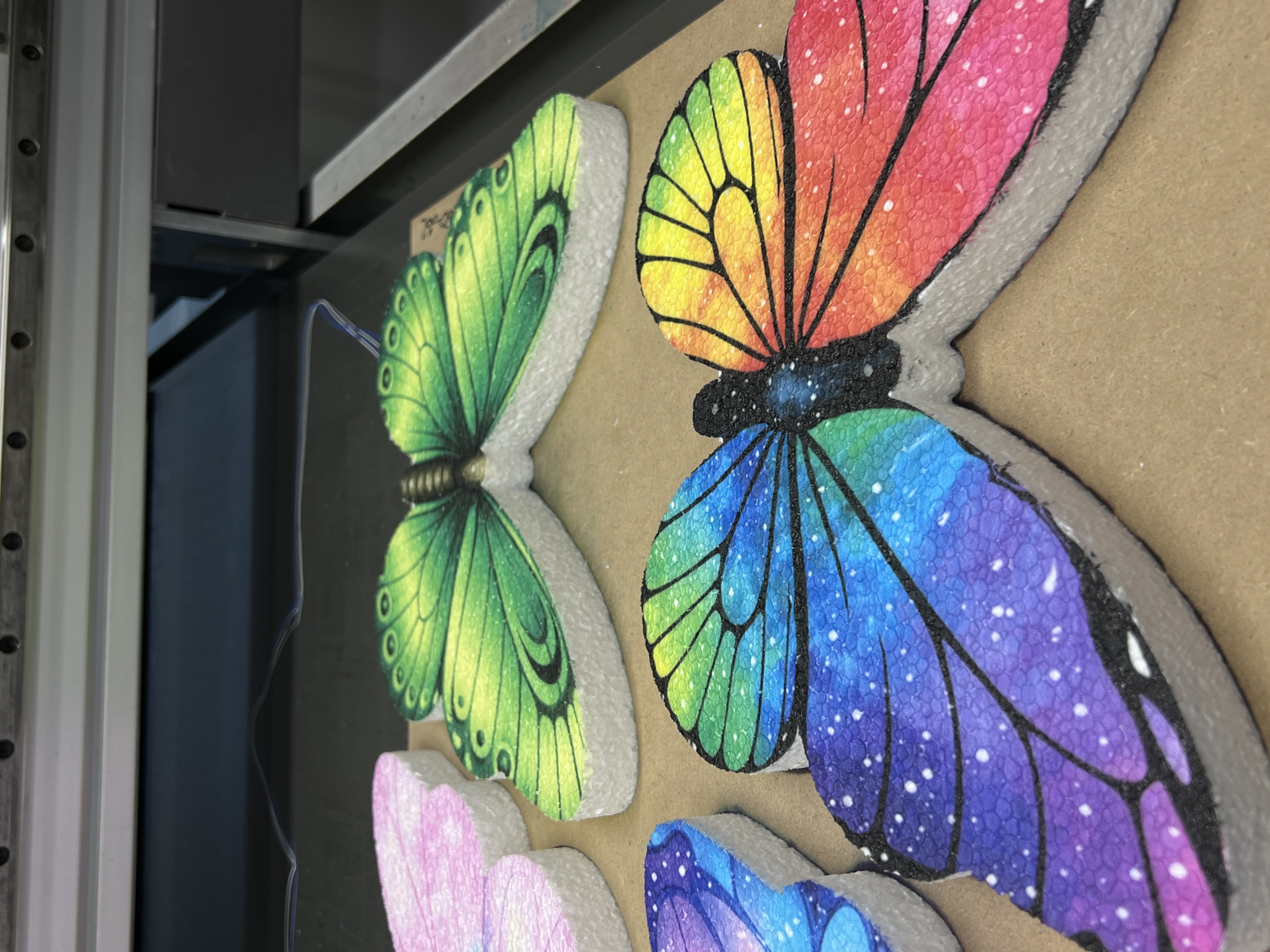 The image captures a close-up, angled view of decorative butterfly cut-outs, likely in a production or display setting.
The primary subjects are several multi-colored butterfly figures, appearing to be crafted from foam board or a similar lightweight material, mounted on a light brown, matte surface. Two butterflies are prominently featured: a vibrant green one on the left and a larger, rainbow-colored one on the right. The green butterfly has detailed veins on its wings and a metallic, dark gold body. The rainbow butterfly's upper wings transition from yellow to orange to red, while its lower wings blend from blue to purple. All visible butterfly wings display a textured, honeycomb-like pattern and are speckled with numerous small white dots, creating a "starry" or "galaxy" effect. The butterflies are three-dimensional, with their white, porous edges of the foam core visible, suggesting they are elevated from the background surface. Parts of a pink and a blue/purple butterfly are also visible at the bottom left and bottom center, respectively.
In the background, particularly to the left and top, industrial-looking machinery or structural components are visible. These include a vertical grey metal beam with circular indentations (possibly bolt heads) and darker grey rectangular sections. A thin, wavy blue wire or tube runs horizontally across the mid-left portion of the frame, passing near the green butterfly.
Above the green butterfly, on the brown mounting surface, there is a small, light-colored square with faint, handwritten dark text that appears to read "9th B." or "9.00". The scene suggests an indoor environment, possibly a workshop or manufacturing facility where these decorative items are being created or prepared for display. There are no specific cues for time of day, weather, or human emotions.