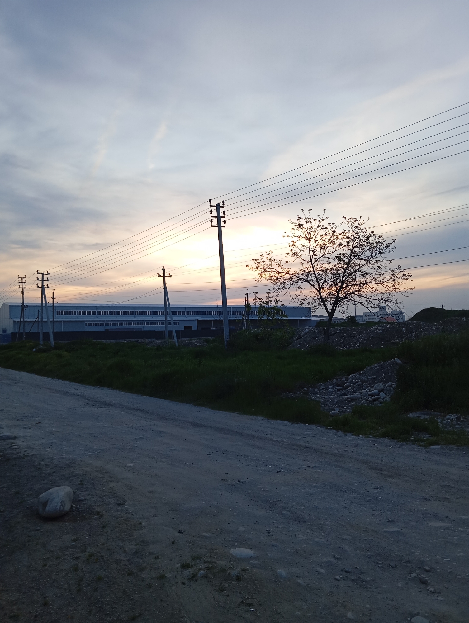 The image depicts a landscape in or near Baksan, Russia, at dusk. An unpaved road dominates the foreground, curving to the right and receding into the distance. A large, light-colored rock sits on the left side of the road. To the right of the road, there is a grassy area interspersed with piles of rocks and dirt.
In the middle ground, a row of electrical poles with multiple wires stretches across the scene, appearing to run parallel to a large, rectangular building with numerous windows, which suggests it could be an industrial or commercial structure. The building is light in color, contrasting with the darkening sky. A tree, silhouetted against the setting sun, is positioned to the right of the electrical poles, and a small cluster of greenery can be seen between the tree and the building.
The sky shows a mix of light and dark clouds, with the sun positioned in the middle, casting a soft, orange glow. The light is diffused, suggesting that the sun is near the horizon. The overall scene conveys a sense of rural or industrial outskirts, captured in the evening light.