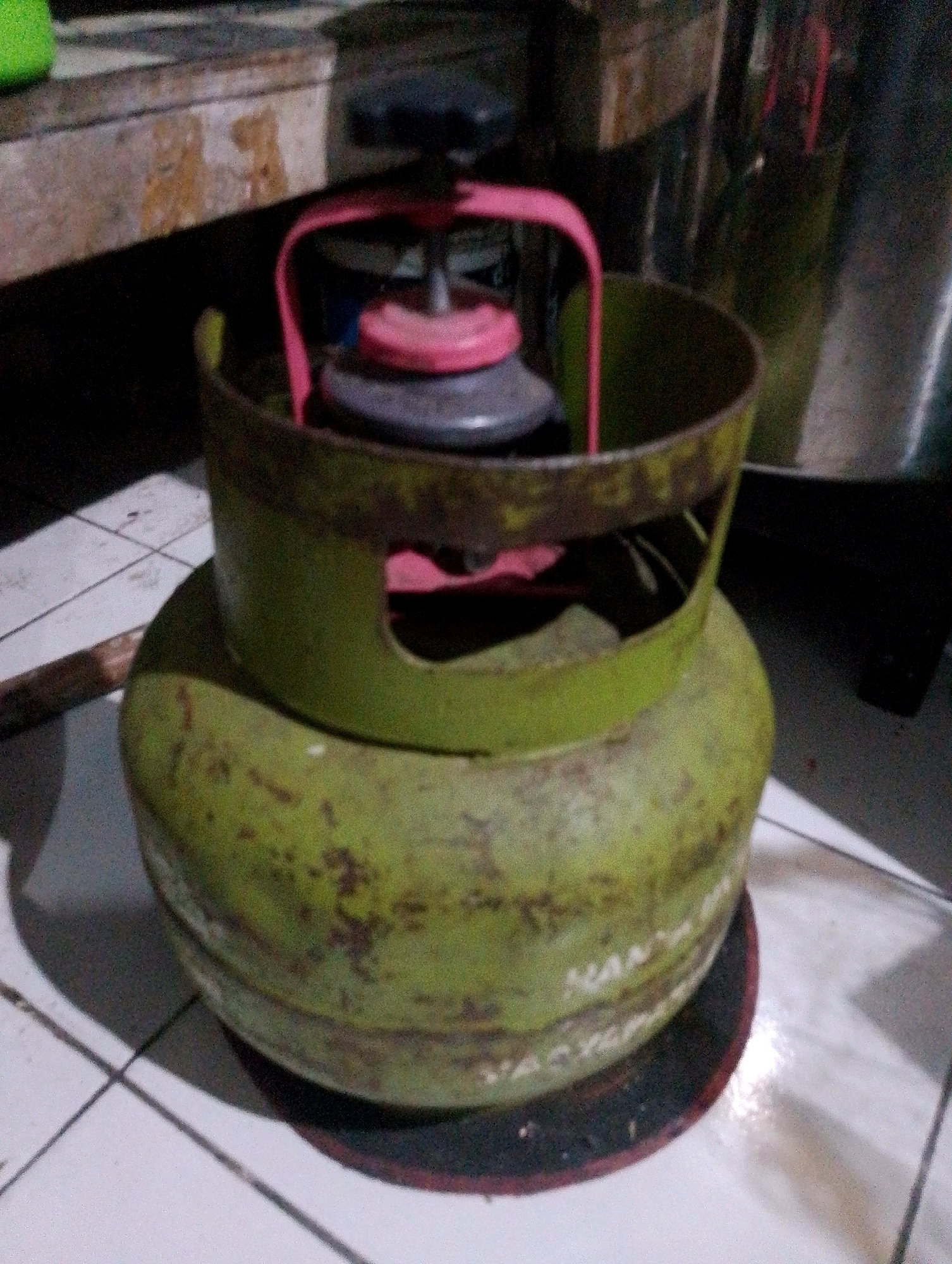The image shows a green propane gas cylinder with a red regulator cap resting on a circular black mat. The cylinder appears to be made of metal and has some scuff marks and discoloration. The regulator cap is a bright pinkish-red color. The cylinder is placed on a tiled floor that is white with gray grout lines. In the background, there is a blurry wooden surface and some metallic objects. The scene is indoors, likely in a domestic setting within South Jakarta, Indonesia. No people are visible in the image.