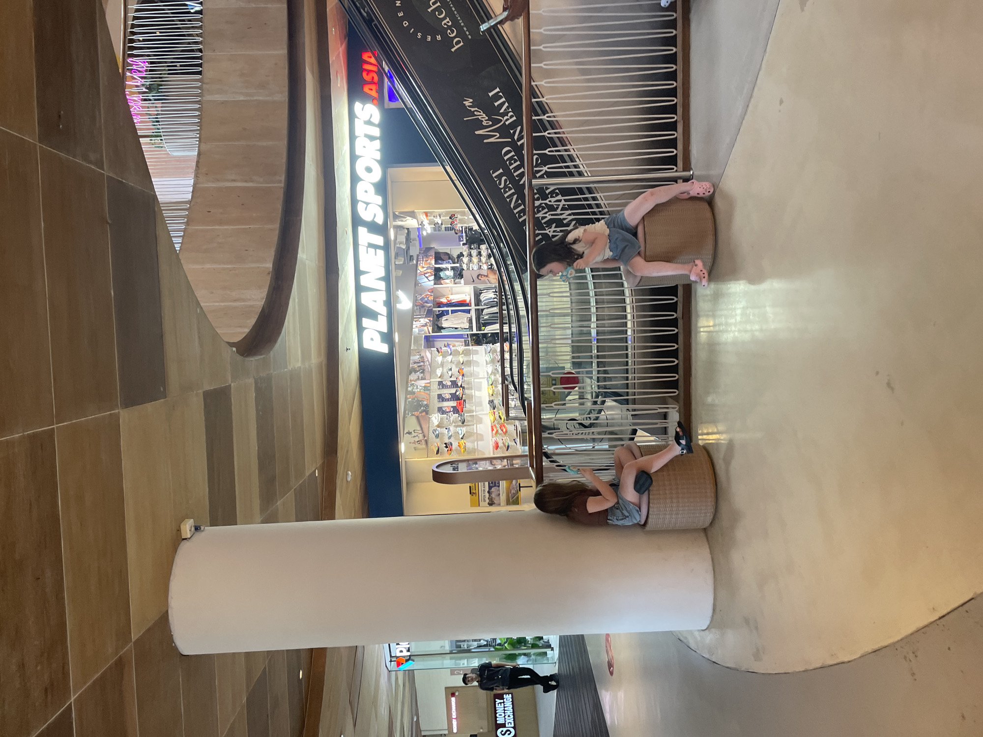 Here is a description of the image:
The image shows an indoor space, possibly a shopping mall or a large commercial building, featuring two women seated on wicker ottomans. The women are positioned in the foreground, each engrossed in a mobile phone. One woman is wearing blue jean shorts and has pink sandals while the other has blue and black sandals and a black knee brace. Both are facing opposite directions and are wearing t-shirts.
In the background is a large, cylindrical white pillar. An escalator ascends upwards behind the women and a storefront for Planet Sports Asia is visible, indicating this may be in Asia. Below the pillar is signage that indicates a currency exchange is located near there. The lighting is bright, and the scene conveys a casual, relaxed atmosphere.