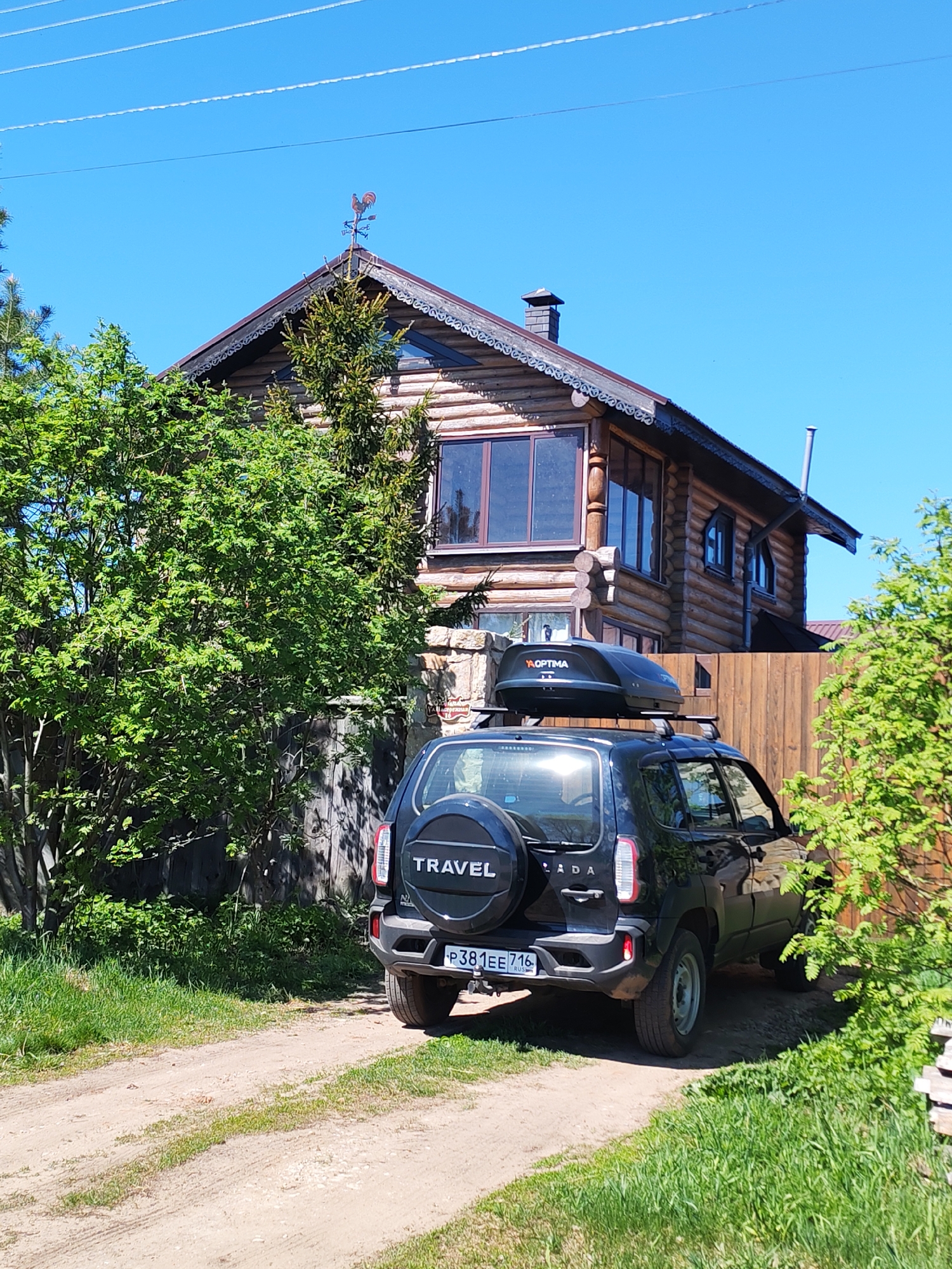 A black Lada Niva Travel car, with a roof box and a spare tire mounted on the back, is parked in front of a log cabin house. The car's license plate reads "P381EE 716 RUS". The house has a decorative roof trim and a weather vane on top. The setting is a rural area, with dirt roads and trees surrounding the house. The sky is clear and blue. The location is presumably in or near Икское Устье, Russia, based on the license plate information.