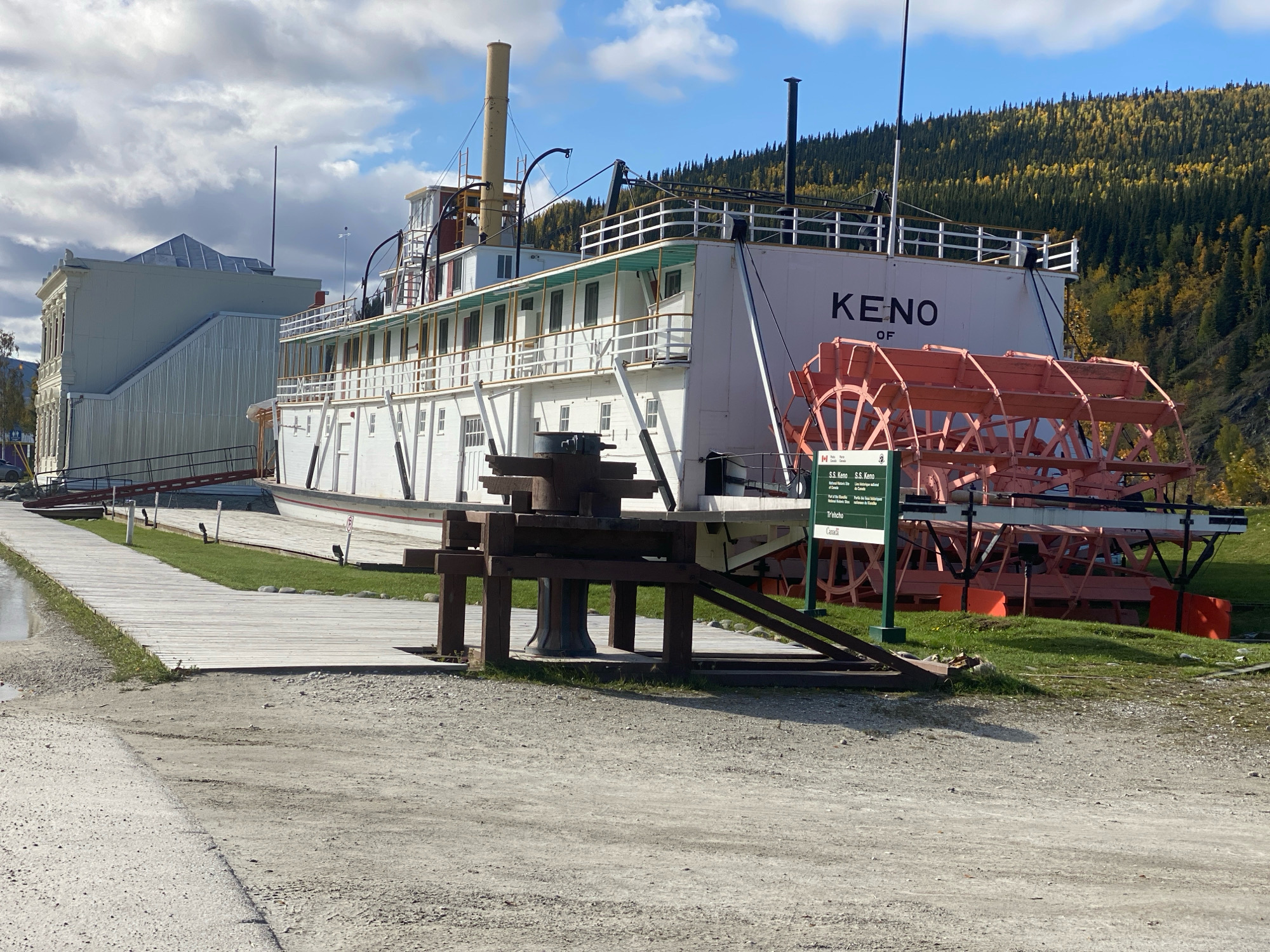 In Dawson City, Canada, the SS Keno, a historical sternwheeler, is moored on land as a museum. The vessel is white with a green trim and features a large, orange paddlewheel at its stern. A wooden boardwalk leads towards the ship, with a ramp for access. Adjacent to the SS Keno is a modern, grey building with a distinctive angular roof. In the background, a forested hillside displays autumn foliage in shades of yellow and green under a partially cloudy sky. Several informative signs are visible near the paddlewheel, including one with the text "S.S. Keno." The ground in the foreground consists of gravel and dirt.