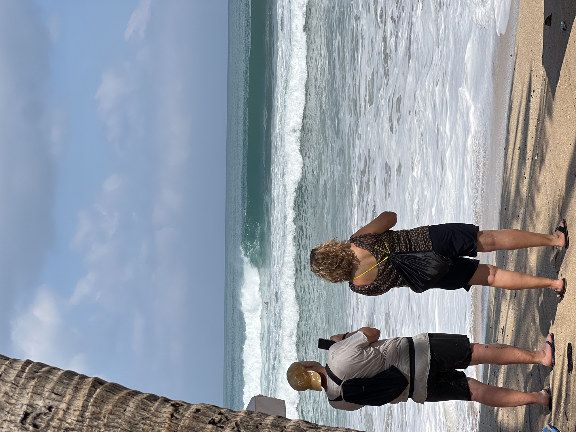 Two people stand on a sandy beach in Kuta, Indonesia, facing the ocean. The person on the left, a male wearing a light-colored t-shirt, black shorts, a yellow baseball cap, and a black backpack, holds a smartphone in his right hand, appearing to take a photo or video. The person on the right, a female with blonde curly hair, wears a patterned top, black shorts, and a black drawstring bag slung over her left shoulder. Both individuals are wearing flip-flops.
In the distance, the turquoise ocean displays significant waves, with white foam crashing on the shore. A surfer can be seen in the water, positioned in front of a breaking wave. The sky above is partly cloudy, with a muted blue hue. A portion of a large palm tree trunk is visible on the left side of the frame, casting a shadow on the sand. The overall scene captures a moment of observation and leisure by the sea in Kuta.