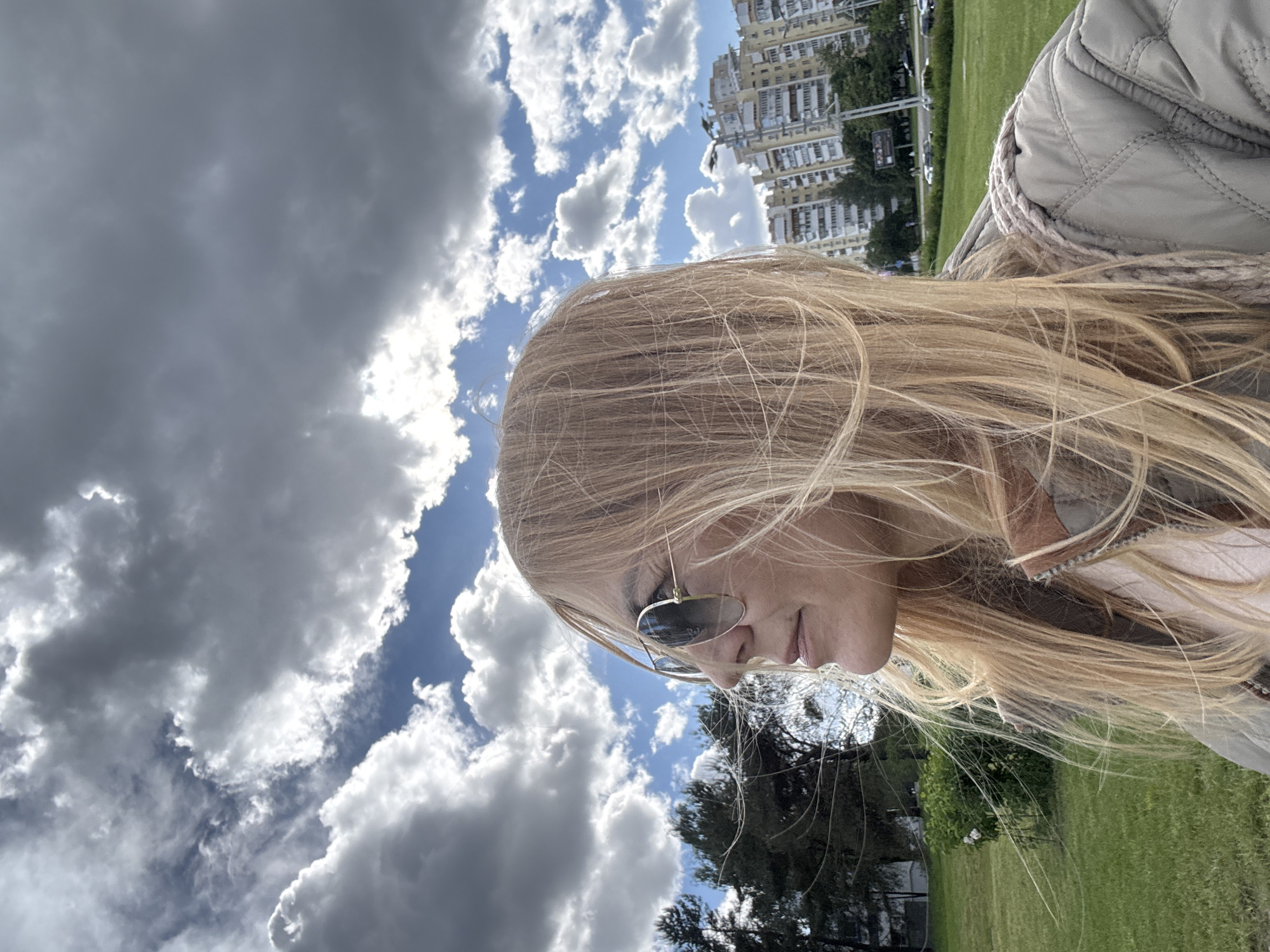 A woman with blonde hair and sunglasses is the primary subject, positioned in front of a partly cloudy sky. Tall buildings are visible in the background, suggesting an urban setting, specifically in Minsk, Belarus. Lush green grass indicates the presence of a park or open green space. The woman's beige jacket is partially visible.