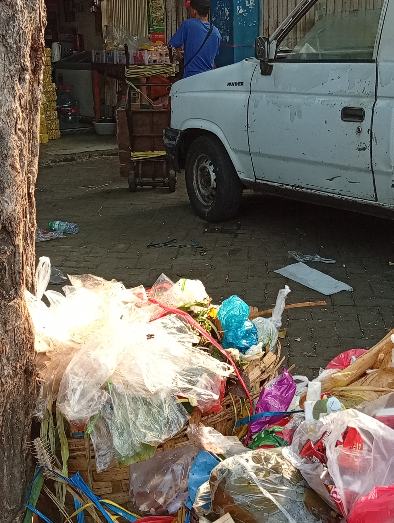 A pile of trash, consisting of plastic bags, bottles, and various debris, is situated on a paved street in front of a storefront. A white pickup truck is parked to the right of the trash pile, with its driver's side facing the viewer. A person wearing a blue shirt and a red cap is partially visible, standing near the truck. The scene appears to be in a commercial area, possibly a market or street stall, with the environment suggesting a developing country setting. The overall impression is one of urban neglect and waste management challenges.