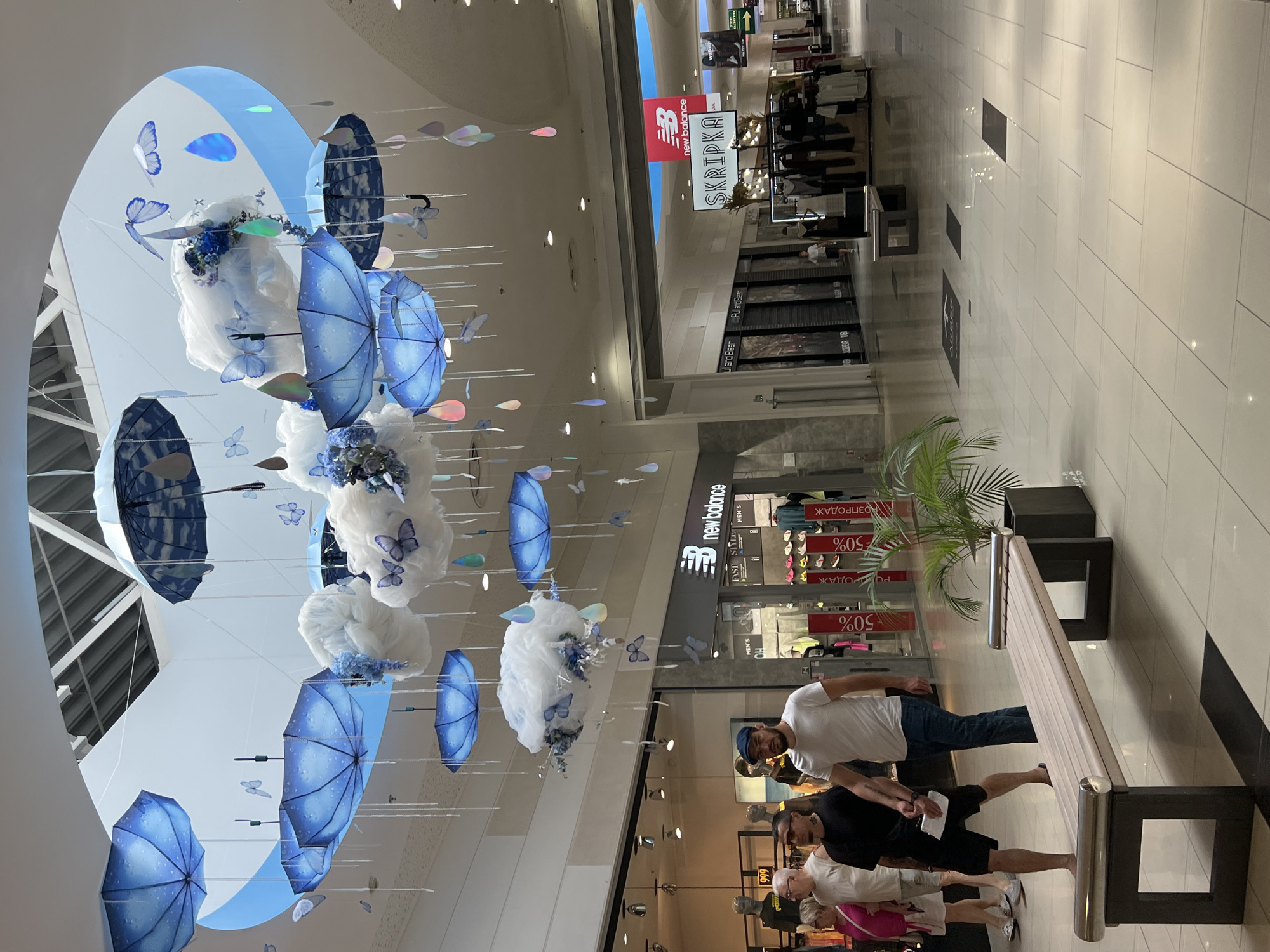 The image shows an indoor shopping mall with a ceiling decoration featuring blue umbrellas, white clouds, butterflies, and hanging teardrop-shaped elements. In the foreground, a light-colored bench is situated on a tiled floor. To the left of the bench, two men and a woman are walking. The man in the center is wearing a white t-shirt and a dark cap, while the man to his left is wearing a black t-shirt and shorts. The woman on the far left is wearing a pink top and white shorts. In the background, various store entrances are visible, including a "New Balance" store and a "SKRIPKA" store. The overall environment is well-lit, suggesting it's daytime. The location is identified as Fontanka, Ukraine.
