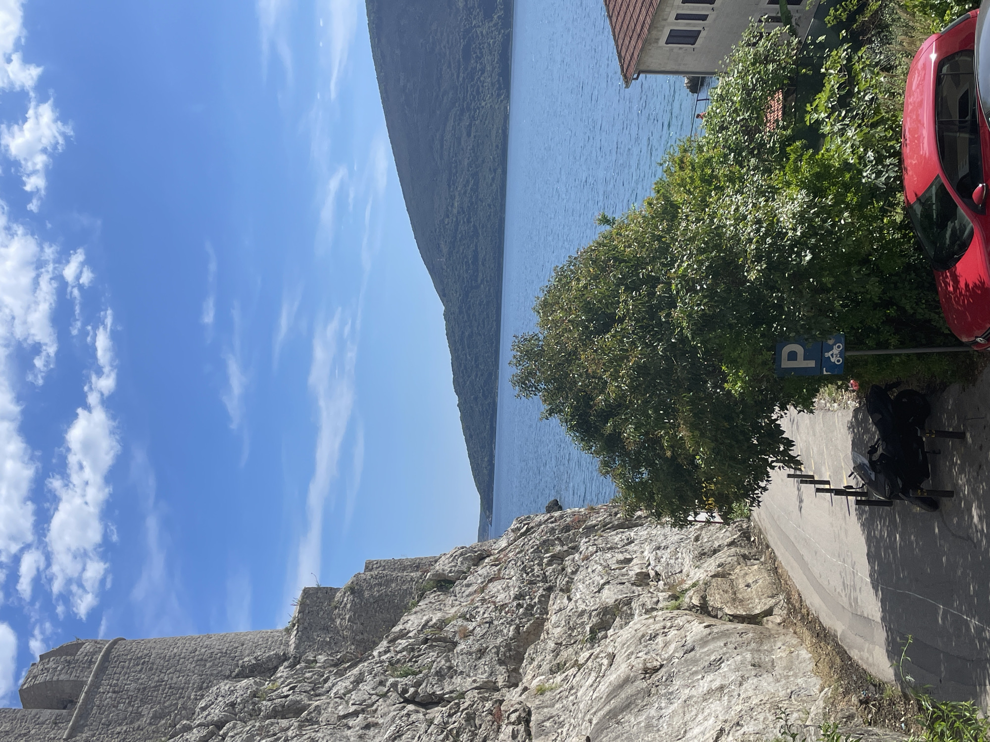 The image shows a view in Herceg Novi, Montenegro, with a stone wall in the foreground leading to a road. The road has a parking sign with bicycle symbol and a parked motorcycle. A tree and red car are near the road. In the background, there is water, a mountain, and a blue sky with some clouds. A building is partially visible in the upper right.