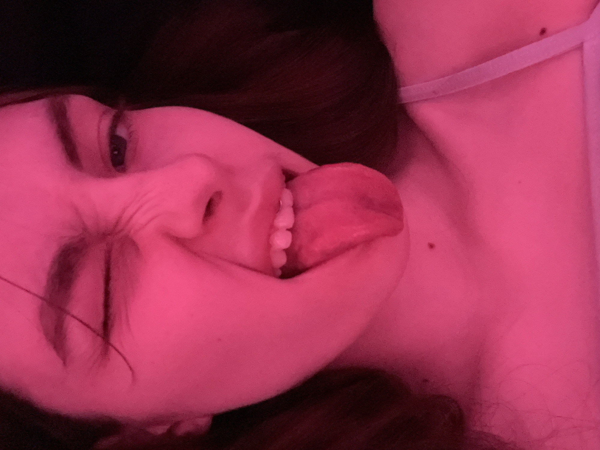 A close-up, low-angle shot shows a person's face distorted into a playful grimace. The person has fair skin, brown hair, and appears to be sticking their tongue out. The overall lighting is a saturated pink hue, possibly due to artificial lighting. The person's right eye is squinted shut, while the left eye is open. Part of the person's neck and shoulder are visible, with a thin white strap suggesting clothing. Dark background elements are also visible. No information suggests this image was taken in St. Petersburg.
