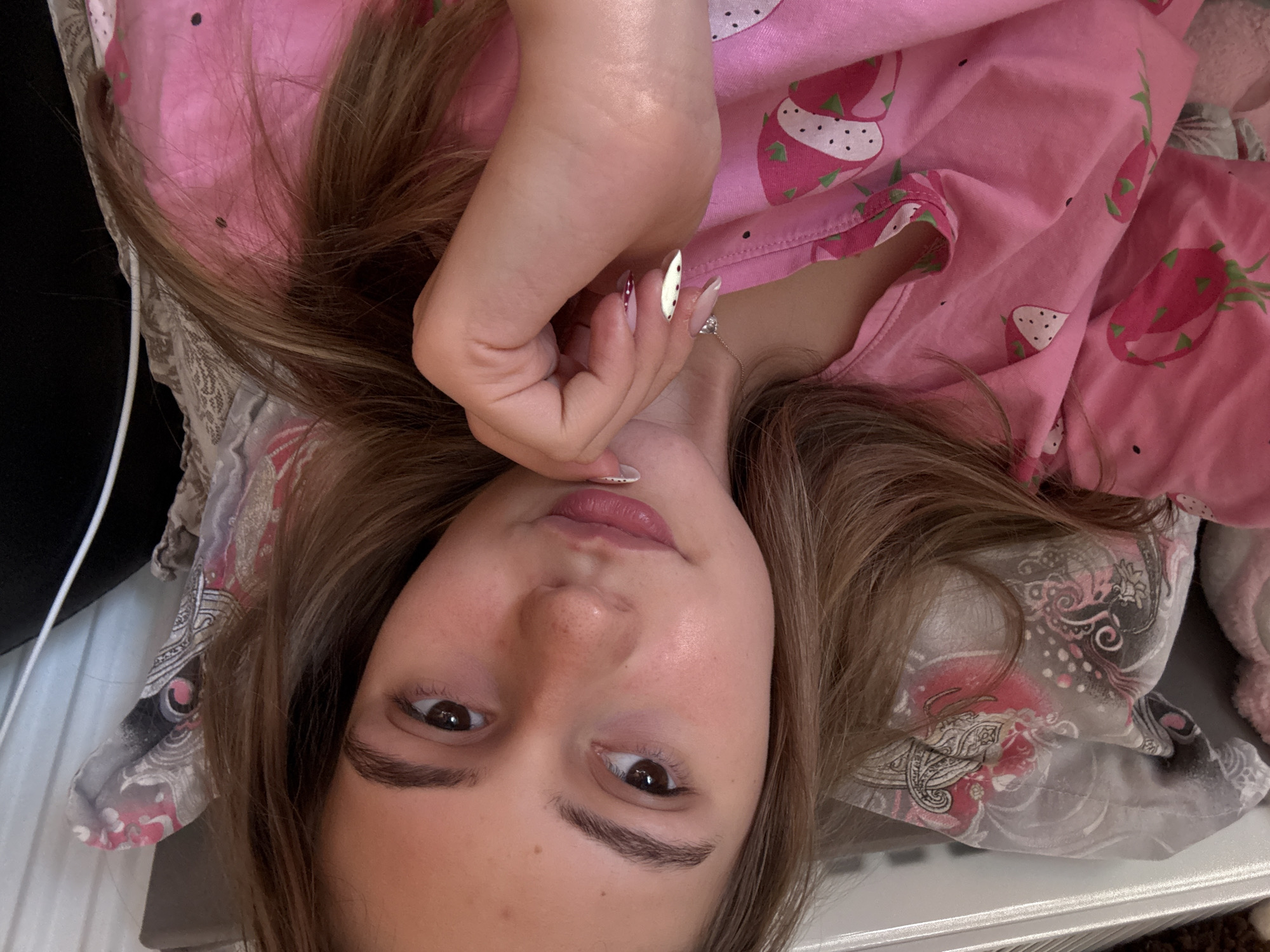 A young woman lies on her back, seemingly on a bed, facing the camera. She is wearing pink pajamas with a strawberry pattern. Her light brown hair is spread around her head on a pillow with a paisley-like pattern. Her left hand is raised and positioned near her face, showcasing long, decorated nails. The background is indistinct but suggests an indoor setting. There is no indication of Panagyurishte, Bulgaria.