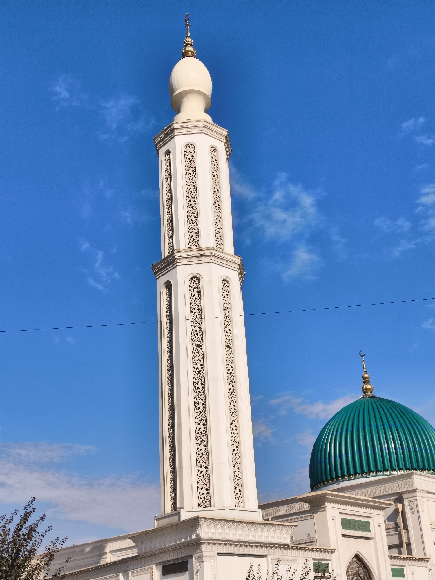A low-angle, full outdoor shot captures the exterior of a mosque under a bright blue sky with scattered white clouds. The main subject is a tall, white minaret with ornate decorative carvings that rises on the left side of the frame. The minaret is topped with a golden finial featuring a crescent moon. To the right of the minaret, a vibrant teal-green dome of the mosque is visible, also adorned with a golden crescent moon at its peak. The mosque structure itself appears to be white with intricate architectural details. A single thin black wire is stretched horizontally across the middle of the sky. In the bottom left corner, the branches of a dark-leaved tree are partially visible, suggesting a natural element near the building. The scene depicts a religious structure in an urban or semi-urban setting, likely Buz, Uzbekistan.