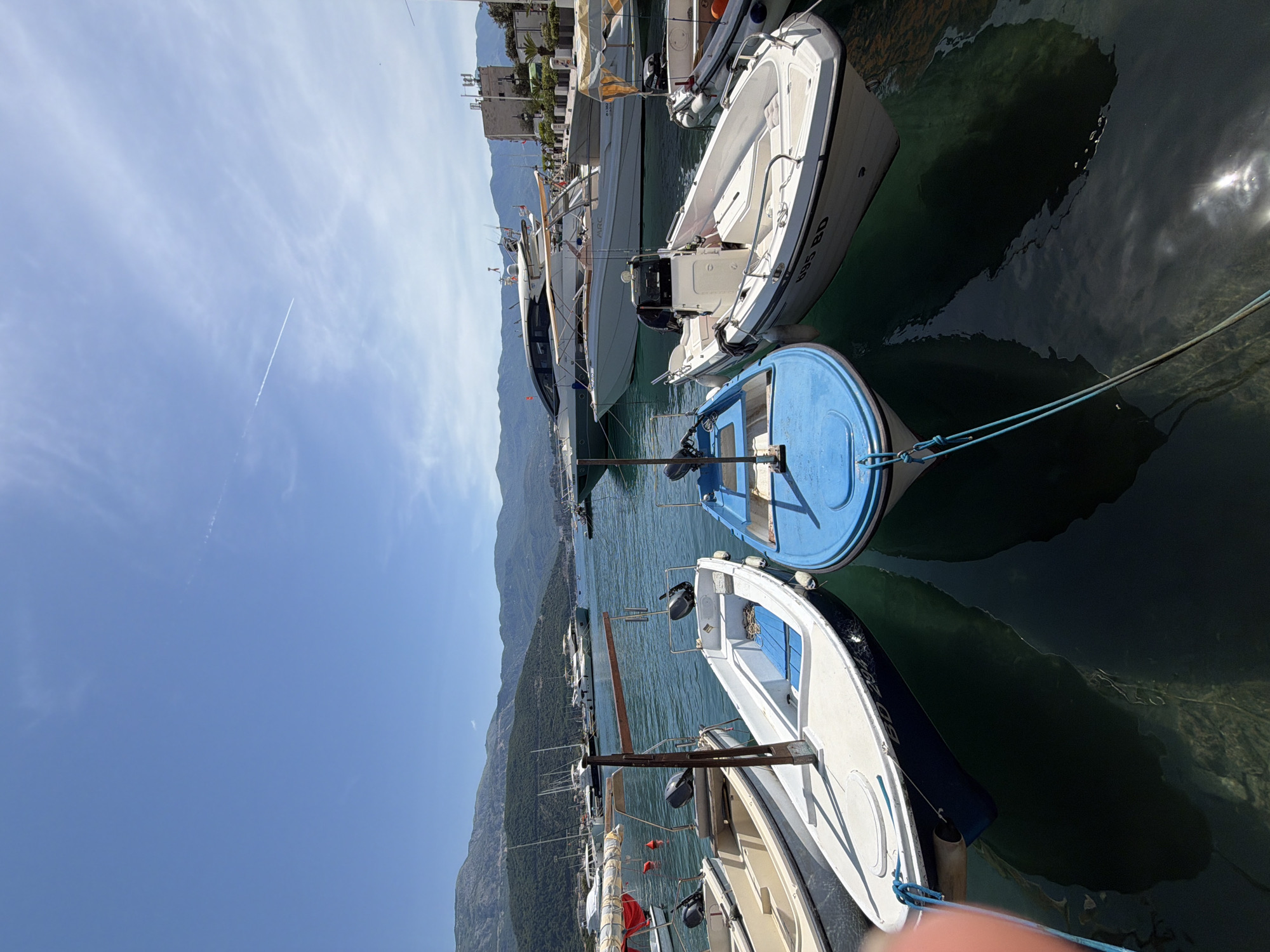 The image shows a waterfront scene in Budva, Montenegro, featuring several boats docked in a harbor. The boats vary in size and type, including small motorboats and larger yachts. The water is relatively calm with a greenish tint. The background includes mountains and some buildings on the shoreline. The sky is blue with a few clouds. There are no people visible in the frame.