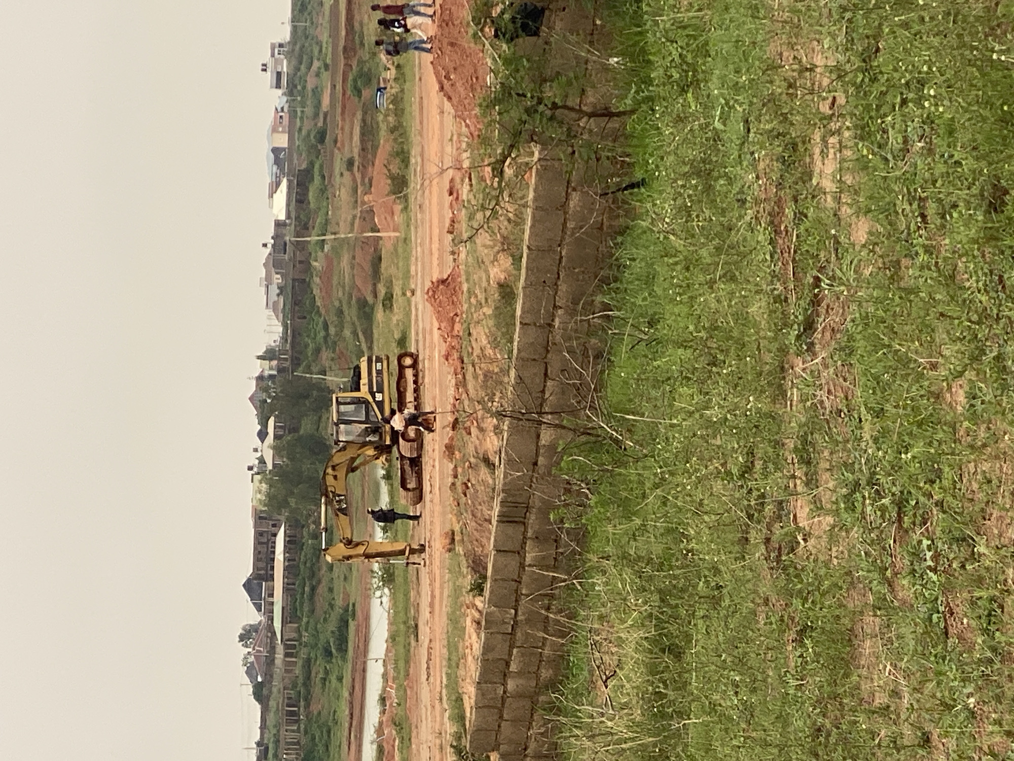 In Kwang, Nigeria, an excavator is positioned on a construction site. Two individuals are standing near the excavator, with one appearing to be interacting with it. Further in the distance, three people are walking away from the site. The area is characterized by cleared land, mounds of dirt, and unfinished buildings in the background, suggesting ongoing development. A water body is visible to the left of the construction site. The foreground is covered with overgrown green grass and a partially constructed brick wall. The sky is overcast, contributing to a muted lighting condition.