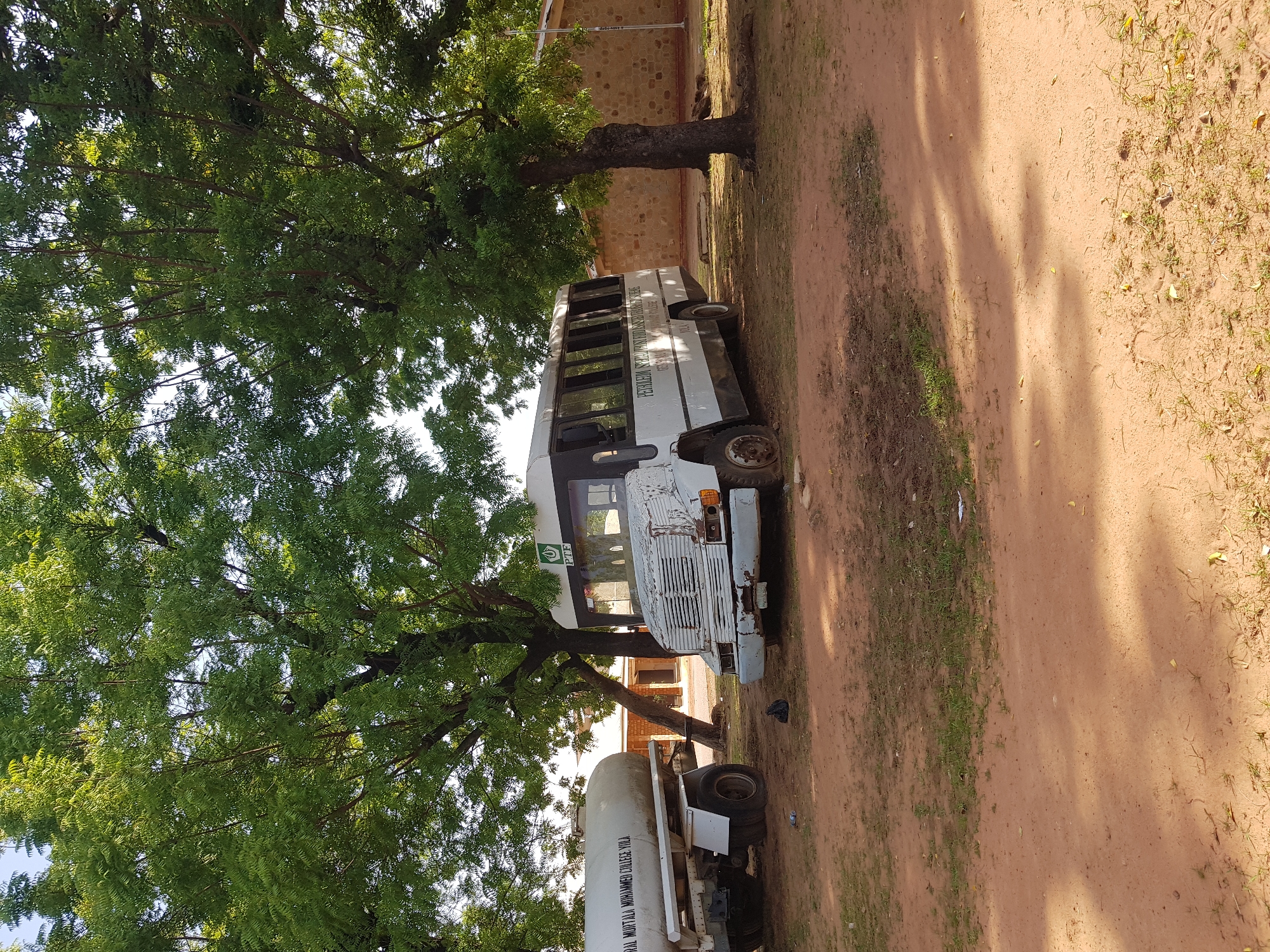 The image shows a white and black bus parked under a large tree with green foliage. The bus is old and appears weathered, with some rust visible on the front. The windows of the bus are dark. To the right of the bus, there is a sandy dirt road with sparse grass. In the background, a building with a stone facade and a red trim is visible. Another vehicle, possibly a water tank truck, is parked behind the bus. The scene appears to be outdoors during the day, with sunlight casting shadows from the tree onto the ground.
Visible text on the bus reads: "PETROLEUM TECHNOLOGY DEVELOPMENT INSTITUTE YOLA". There is also a green logo with white text that is difficult to read. Text on the water tank truck reads: "NGUL MOHTAIJI MOHAMMED JUDEA YOLA".
The overall impression is that of a static scene, possibly at an educational institution or a vocational training center in Jimeta, Nigeria. The time of day appears to be daytime, likely morning or afternoon, given the quality of light and shadows. The weather seems clear.
