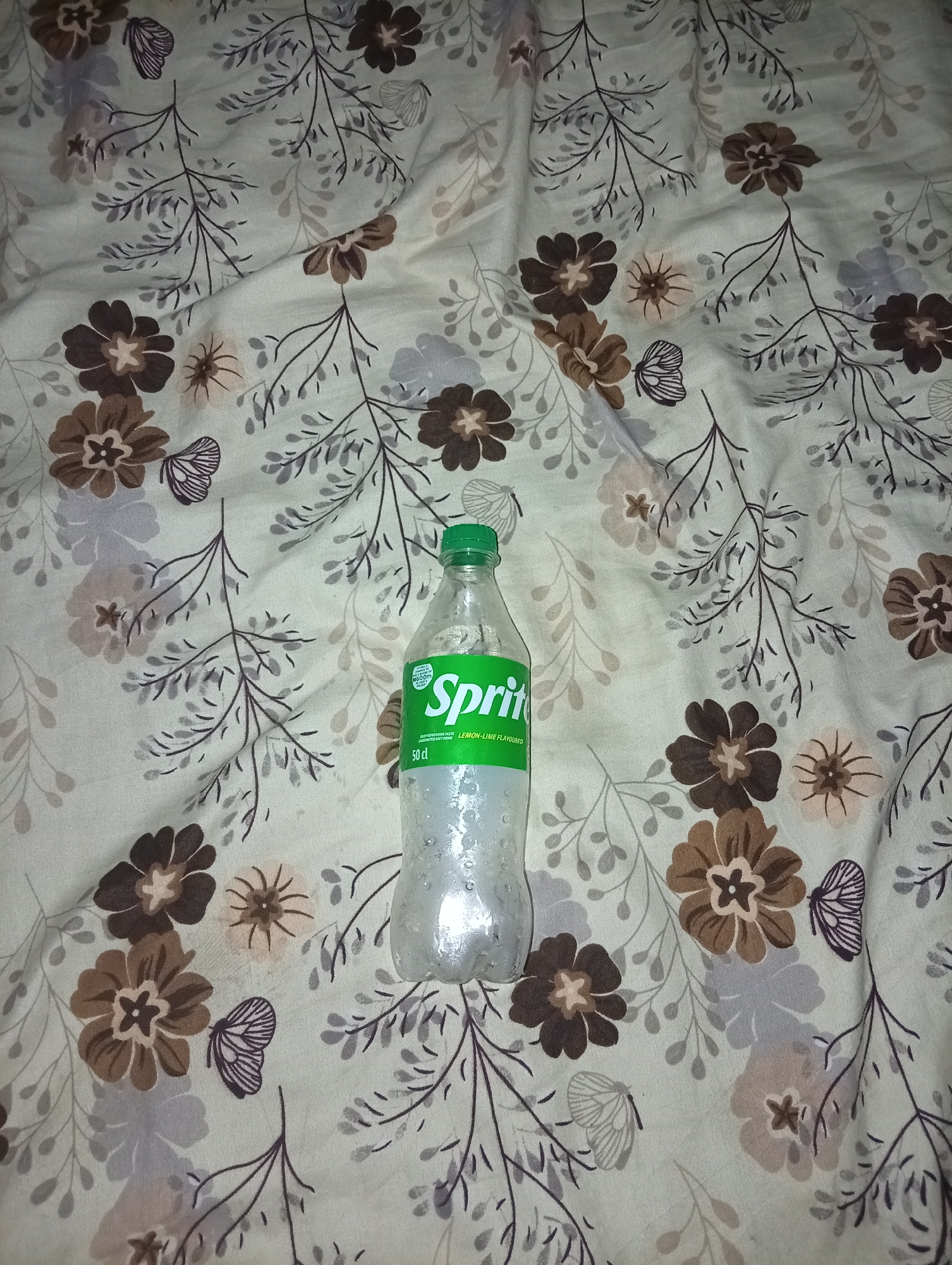 The image depicts a 50 cl plastic bottle of Sprite on a patterned fabric surface. The bottle appears to be slightly frosted. The fabric features a floral and butterfly motif in shades of brown, grey, and off-white. The location is Unknown, Unknown.