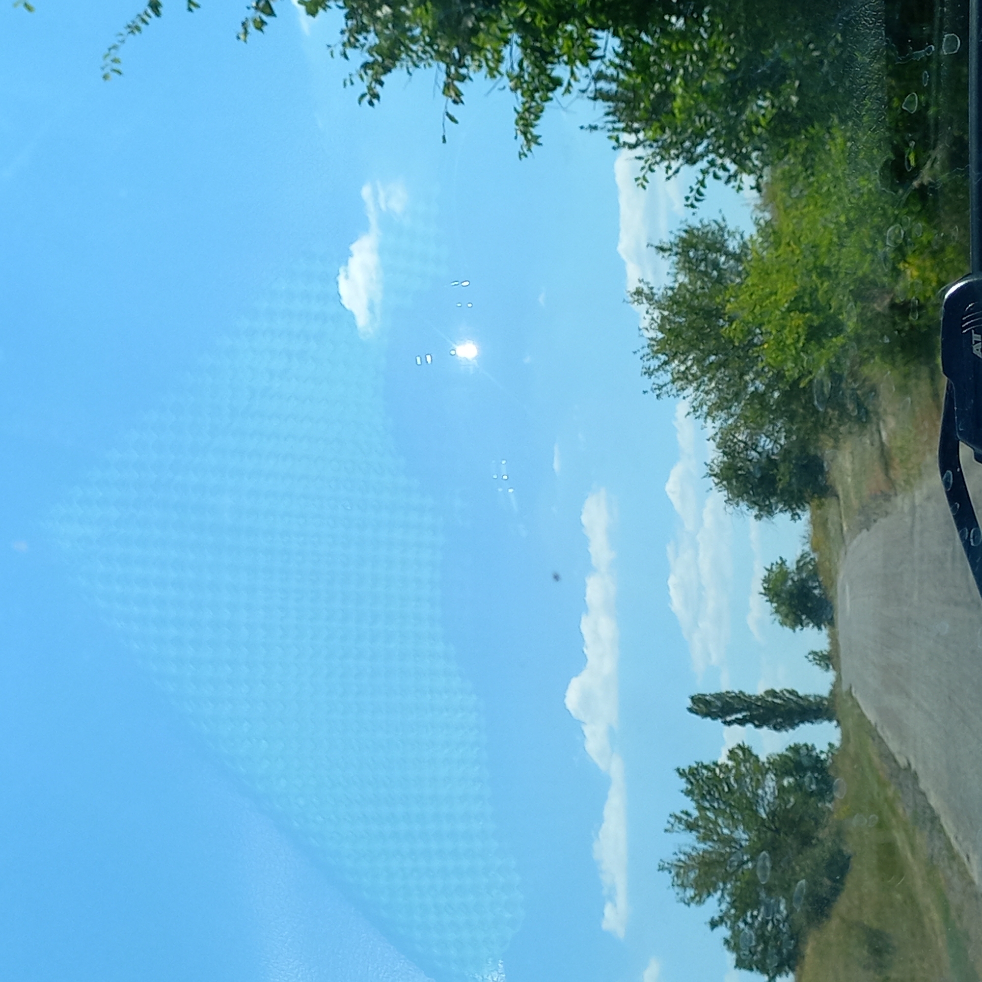 The image captures a view through a windshield, showing a road winding uphill through a rural landscape under a bright blue sky with scattered white clouds. The scene appears to be taken from a vehicle, as suggested by the presence of the car's dashboard in the lower part of the frame and reflections on the glass. The surrounding vegetation includes green trees and bushes, with a tall, slender tree standing out to the left of the road. The sun is visible high in the sky, causing glare and lens flares on the windshield, with some circular water spots or smudges also present. There are no people or specific landmarks that would indicate the city of Klyntsi, Ukraine.
