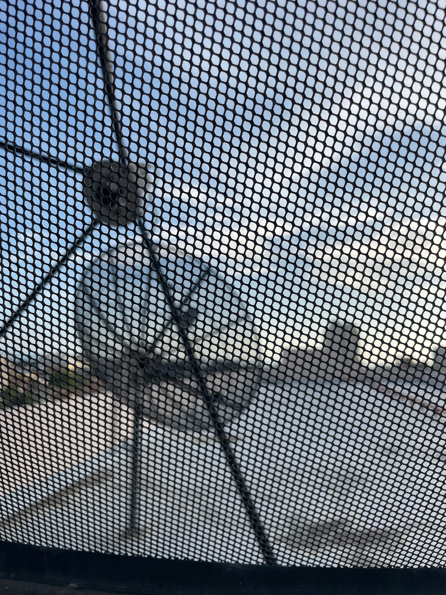 An outdoor, eye-level, close-up, vertical shot captures a black mesh screen obstructing a view of a satellite dish and the sky in Pattaya City, Thailand. The mesh screen, composed of small, uniform hexagonal holes, dominates the foreground and creates a patterned overlay on the scene. Through the mesh, a portion of a satellite dish is visible, along with its supporting arm, in the upper left quadrant of the image. The dish appears to be dark in color. Beyond the dish and mesh, a pale blue sky with diffused white clouds fills the background. In the middle ground, there are indistinct shapes that suggest buildings or structures, rendered blurry by the mesh and distance. The lower portion of the image features a dark, solid band, likely the edge of the screen or a framing element. The lighting suggests it is daytime, with the sun likely behind the camera or diffused by clouds.