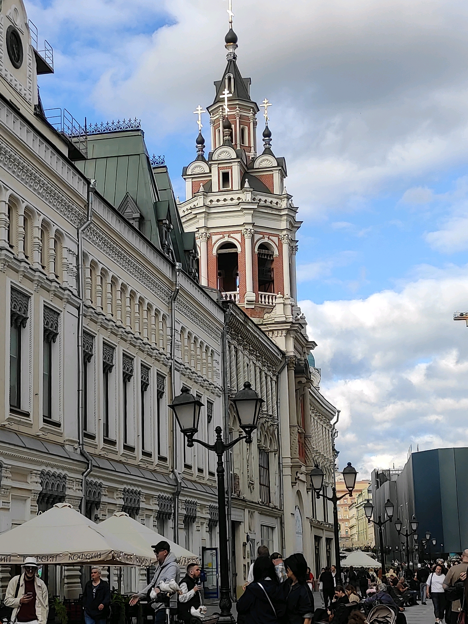 An eye-level, medium shot captures an ornate, white and red bell tower on the right side of the image, piercing a cloudy blue sky. The tower features arched windows and multiple levels, topped with a dark, domed spire adorned with crosses. To the left of the tower, a multi-story building with elaborate white facade detailing and numerous arched windows stretches into the distance. Several dark street lamps are visible along the sidewalk. In the foreground, several people are casually walking and standing. Some are under white umbrellas that appear to be part of a cafe or market. The scene suggests a bustling urban environment, possibly a city street with historical architecture.