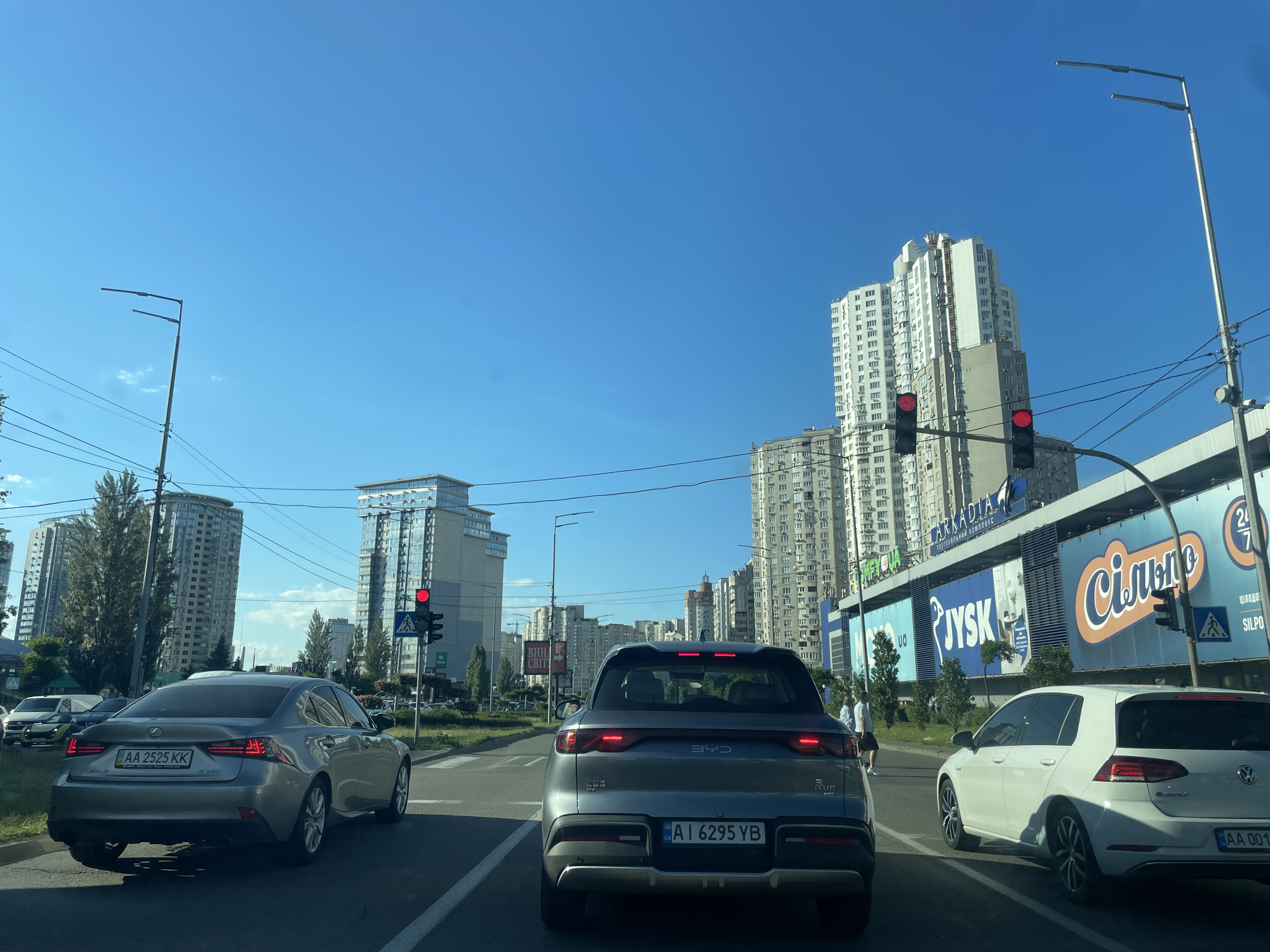 On a clear, sunny day in Kyiv, Ukraine, traffic moves along a multi-lane road lined with modern apartment buildings and commercial establishments. The perspective is from within a vehicle, looking forward. In the foreground, a grey BYD Seal electric car is in the center lane, its brake lights illuminated. To its left, a silver Lexus sedan displays the license plate "AA 2525 KK". To the right, a white Volkswagen Golf with license plate "AA 001..." is in the right lane.
In the middle ground, several traffic lights are visible, with at least one red light showing. Pedestrians are present on the sidewalk to the right. Dominating the background are tall, multi-story buildings, including a distinctive white and grey skyscraper with many windows. A large billboard for "Silpo" supermarket and "Jysk" furniture store is prominent on the side of a building. Overhead power lines and streetlights are also visible. The sky is a vibrant, cloudless blue.