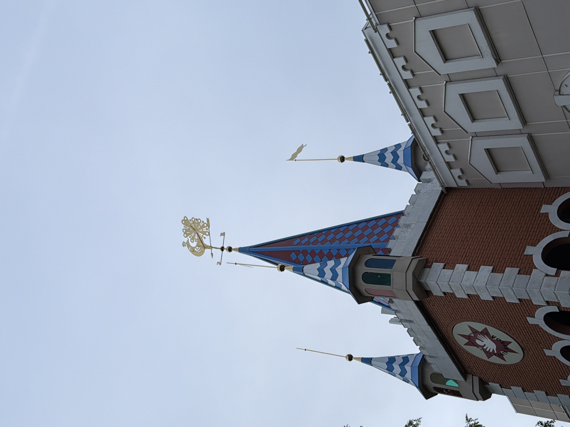 The image depicts the architecture of Sleeping Beauty Castle, specifically the roof details, set against a clear, pale sky. The structure features conical turrets with blue and white zigzag patterns. A taller turret displays red and blue diamond patterns. The castle is constructed with red brick and adorned with white crenellations. A gold weather vane sits atop the tallest turret. At the lower section of the castle's visible structure, a circular emblem is noticeable. The location is in an unknown city.