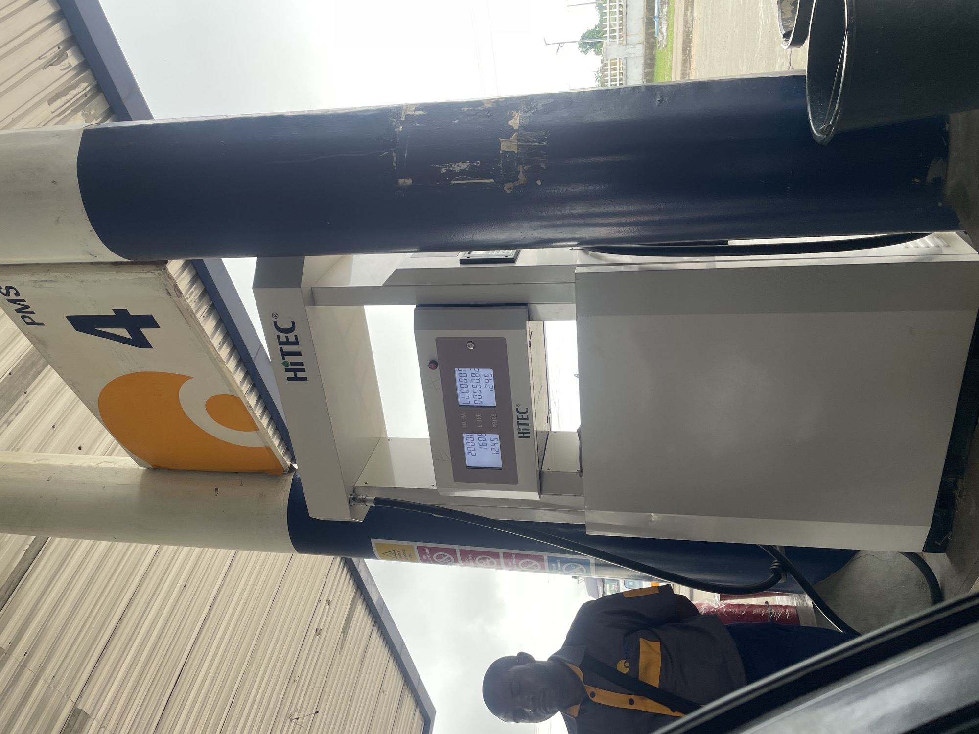 An outdoor scene at a gas station, likely in Karmajiji, Nigeria, features a fuel dispenser with a digital display and a male attendant standing to the left. The fuel dispenser, labeled "HiTEC," shows transaction details including amounts in NAIRA, LTRF, and PRICE. The attendant, wearing a gray and yellow uniform with a black strap across his chest, is looking downwards. The dispenser is situated under a covered area with a ribbed metal roof. Behind the dispenser, a large, dark blue pillar supports the roof structure. In the background, a fence and some vegetation are visible under an overcast sky. Several buckets are placed on the ground near the dispenser.