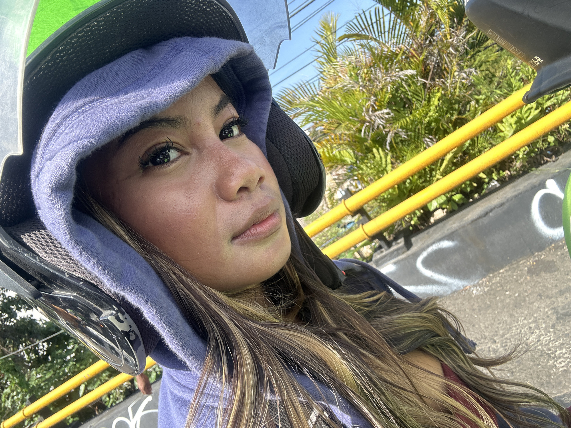 A young woman is taking a selfie while wearing a motorcycle helmet and a hooded jacket. She has long, brown hair with blonde highlights and is looking directly at the camera. The background shows some greenery, yellow poles, and a clear blue sky, suggesting an outdoor setting, possibly in Denpasar, Indonesia, given the context. The image has a slightly tilted, close-up perspective.