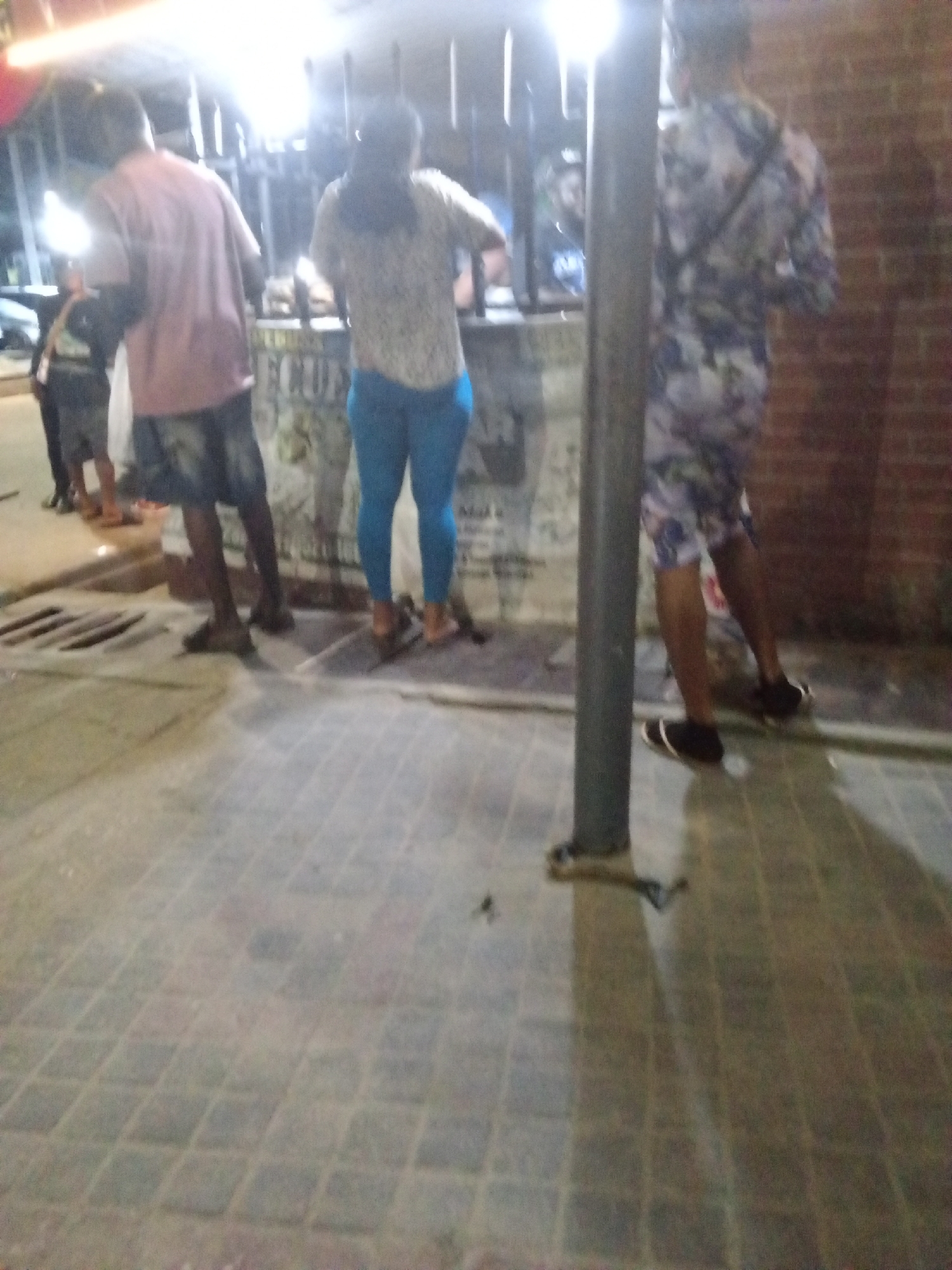 A nighttime scene shows people gathered around a vendor's setup in Ado, Nigeria. The location appears to be outdoors on a paved street. At least four individuals are visible: two men, a woman, and a child. The woman wears light blue leggings and a patterned top. One man wears a pink shirt and dark shorts, while the other wears a patterned shirt and shorts set. The child is dressed in dark clothing. They are standing near a stall or counter that is partially obscured by a vertical post. A brick wall is present in the background on the right side of the image. The ground is paved with rectangular tiles.