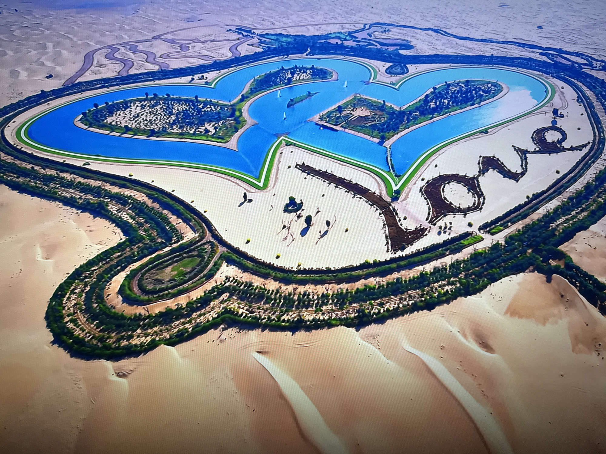 This aerial drone image depicts a heart-shaped lake resort in Burgas, Bulgaria. The resort features two large blue heart-shaped lakes, connected by smaller waterways. The surrounding land is a mix of desert dunes, landscaped green areas with trees and shrubs, and pathways. In the center of the lakes, several small white sailboats are visible. The resort's perimeter is lined with greenery and winding paths. The word "LOVE" is artfully formed with dark landscaping on the right side of the image, adjacent to the heart-shaped lakes. The overall scene conveys a sense of luxury and leisure within a vast, arid environment.