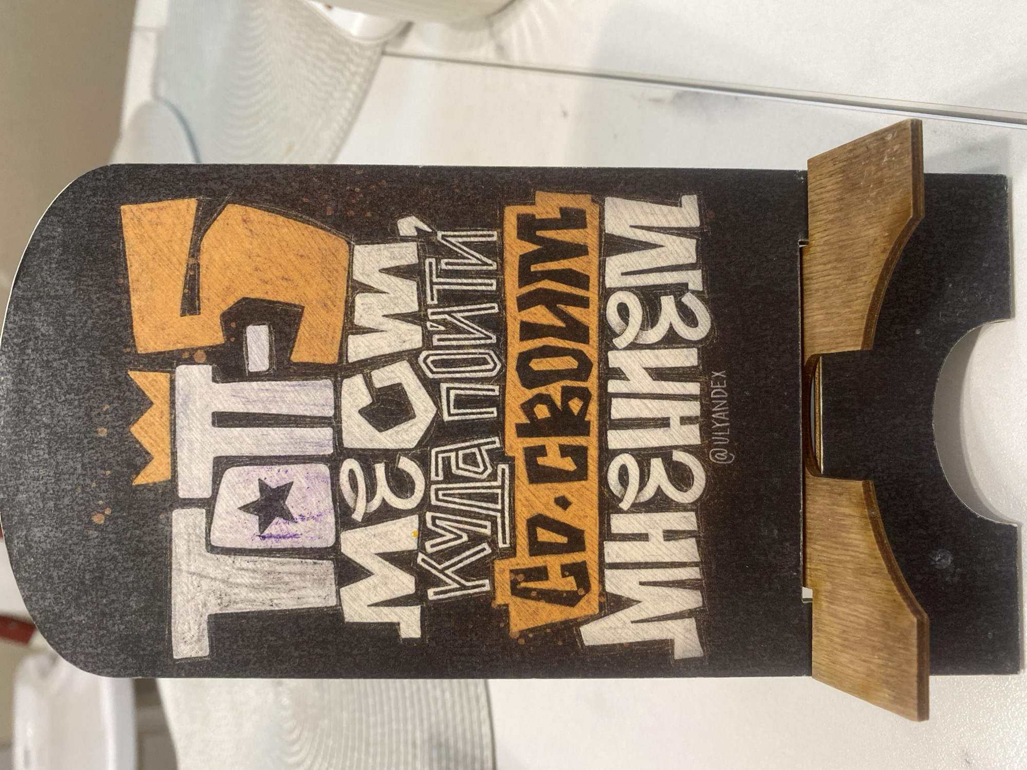 A vertically-oriented close-up shot reveals a black finger skateboard deck adorned with stylized lettering and held upright by a wooden finger skateboard rack. The deck has a rounded top and a flat bottom with cut-outs for the wheels. The artwork features large, stylized, overlapping letters and shapes in white and orange. The lettering reads, in Russian, "ТО, ЧТО МЫ КУДА ПОЙТИ СО СВОИМ МНЕНИЕМ." The skateboard stand is light brown wood. The hashtag "@ULYANDEX" is visible to the right of the skateboard art. The background is white and out of focus. The location is not identifiable.
