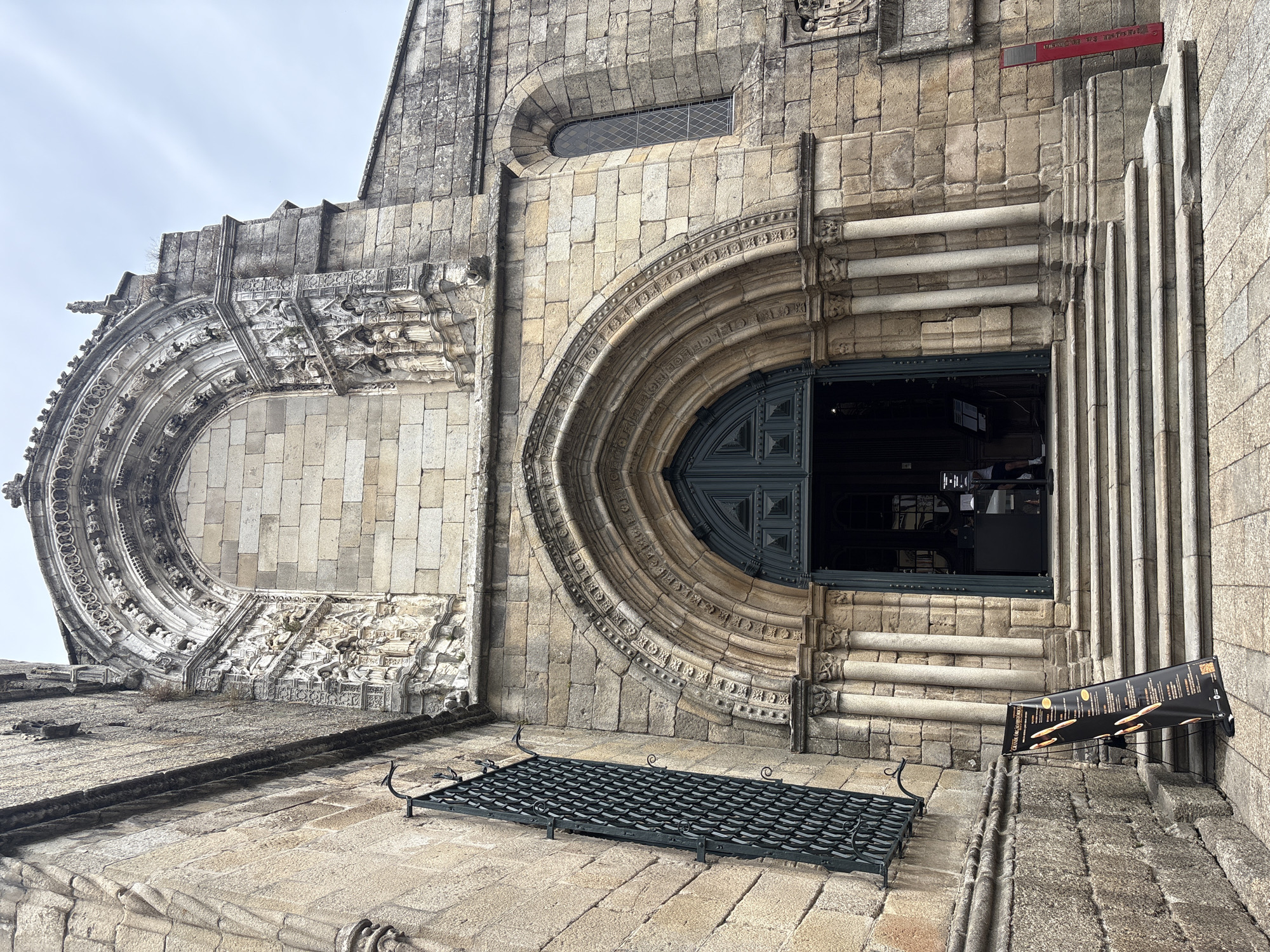 The image shows the entrance to the Church of São Francisco in Guimarães, Portugal. The church is constructed of stone and features a large arched doorway with double doors and elaborate stone carvings above. The entrance is accessed by a wide set of stone steps. To the left of the entrance, a tall, dark metal grate covers a window. A black banner with yellow text and images stands on an easel to the left of the steps. Inside the doorway, a dark interior is visible, with people standing near a counter. A red marker stands on the right side of the steps. The sky is overcast.