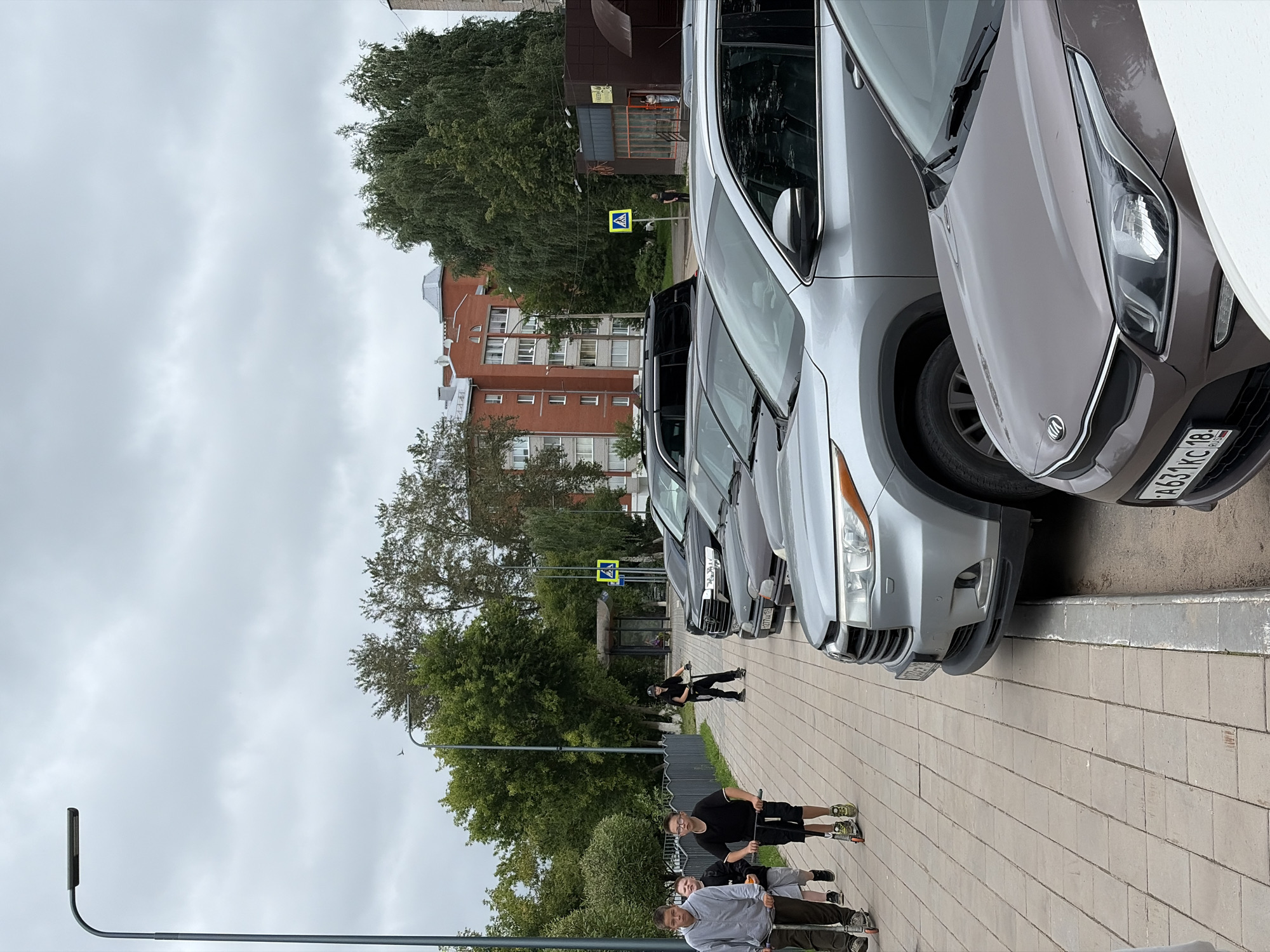The image depicts a cloudy day on a sidewalk in Votkinsk, Russia, with several individuals riding scooters and parked cars. In the foreground, three young males are riding scooters on a paved sidewalk. The person furthest to the left is wearing a grey hooded sweatshirt and dark pants, while the middle person is wearing a black t-shirt and shorts. The third person, further back and to the right, is wearing a black shirt and pants and a helmet. Behind them, a black SUV is parked, followed by a silver SUV, and a brown Kia in the extreme foreground. A pedestrian crossing sign is visible in the background, along with a bus stop shelter. Trees and multi-story brick buildings form the backdrop of the scene.