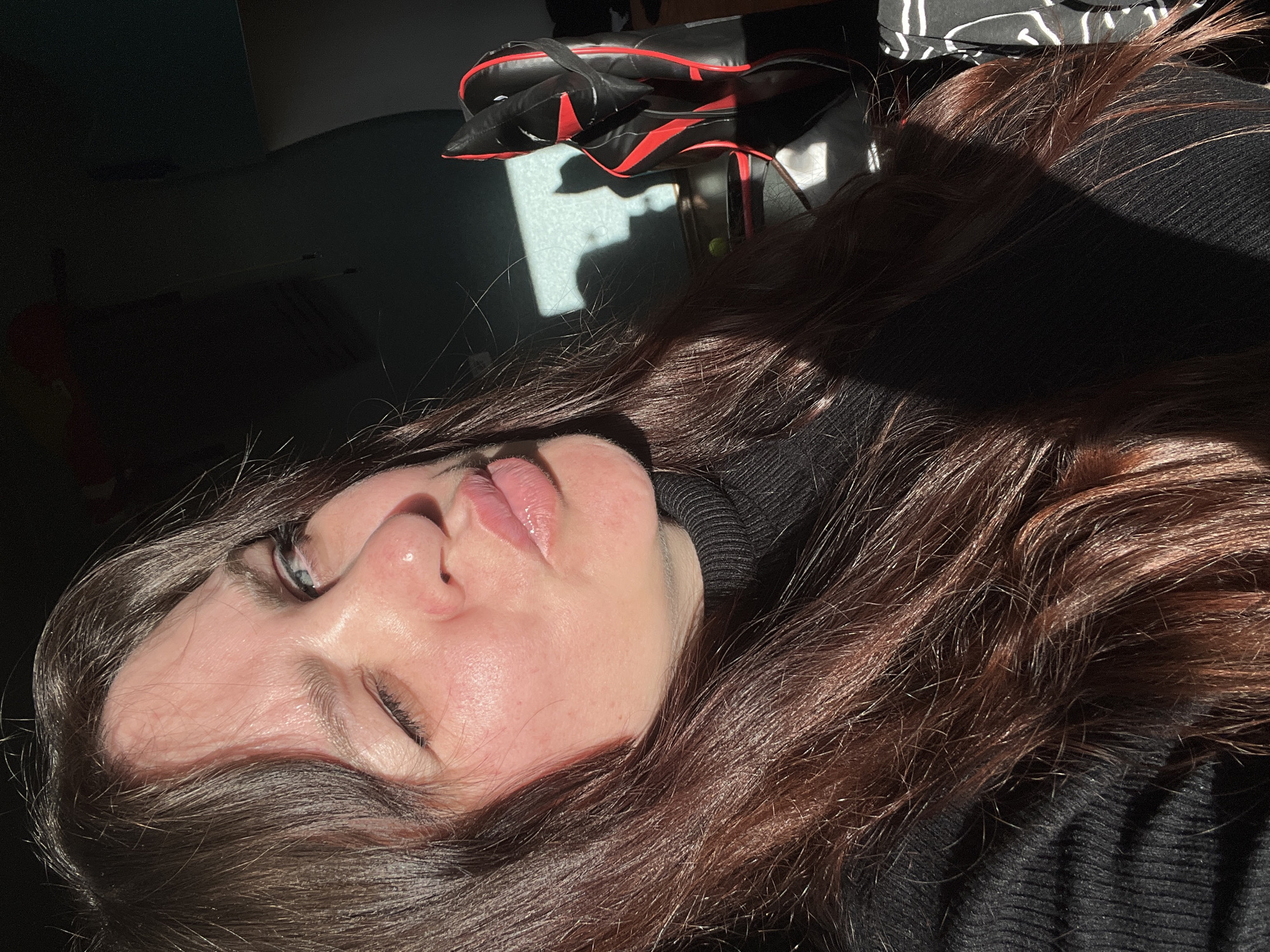A woman with brown hair and fair skin is lying down, illuminated by sunlight. She is wearing a black turtleneck sweater. Beside her head, there is a black bag with red accents. The location in the city of Noviy, Russia is not identifiable from the image.