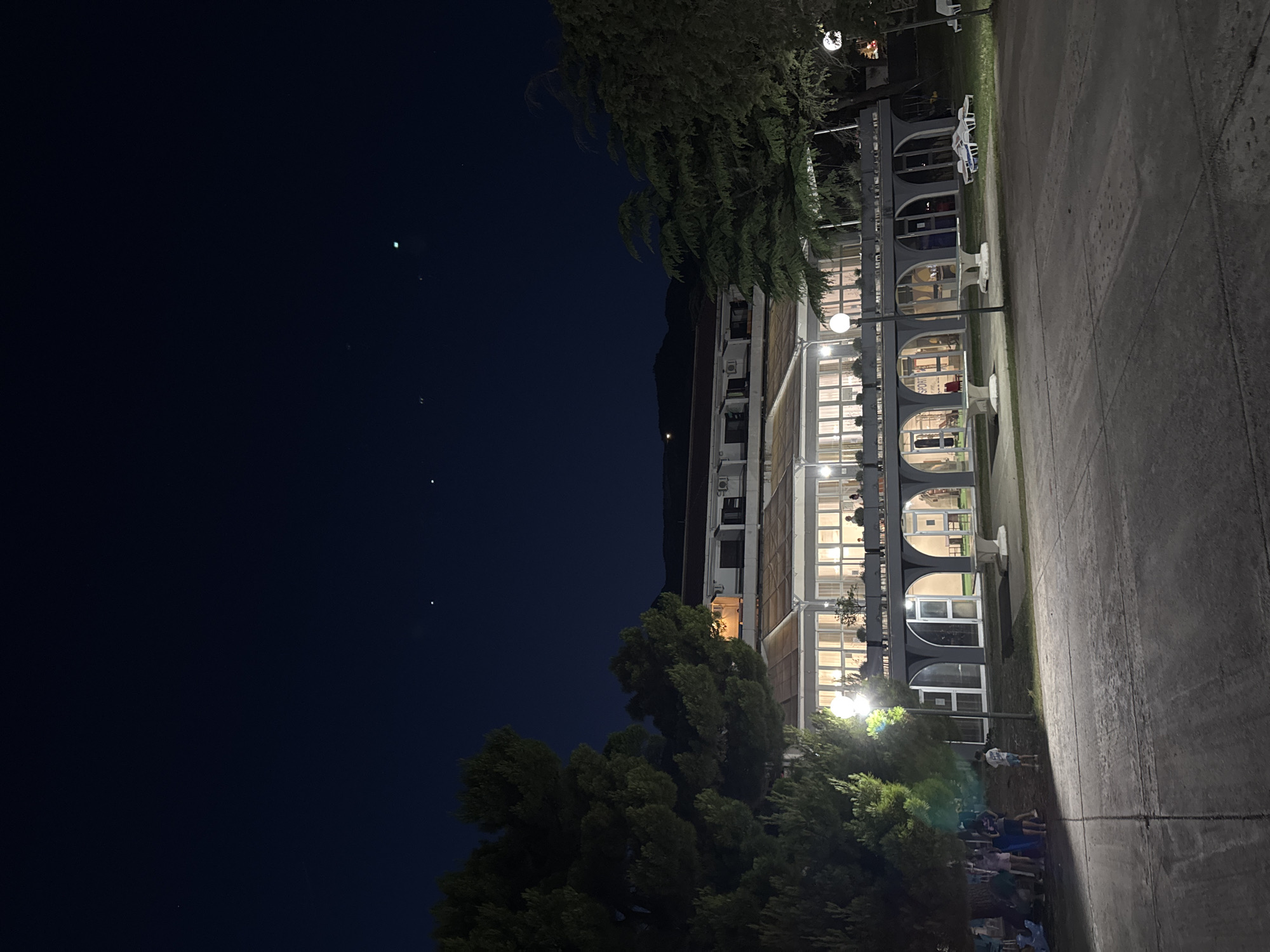 This image depicts the exterior of a building at night in Boreti, Montenegro. The building appears to be a hotel or resort, with a long, multi-story structure featuring numerous arched windows and a glass-enclosed veranda. People are visible inside the building, and some are gathered in front of it, near trees and a streetlamp. The ground in front of the building is paved, and to the right, there is a grassy area with lounge chairs and a distant light. The sky above is dark blue, with several small points of light visible, possibly stars or distant lights. The overall scene suggests a relaxed evening at a coastal resort.