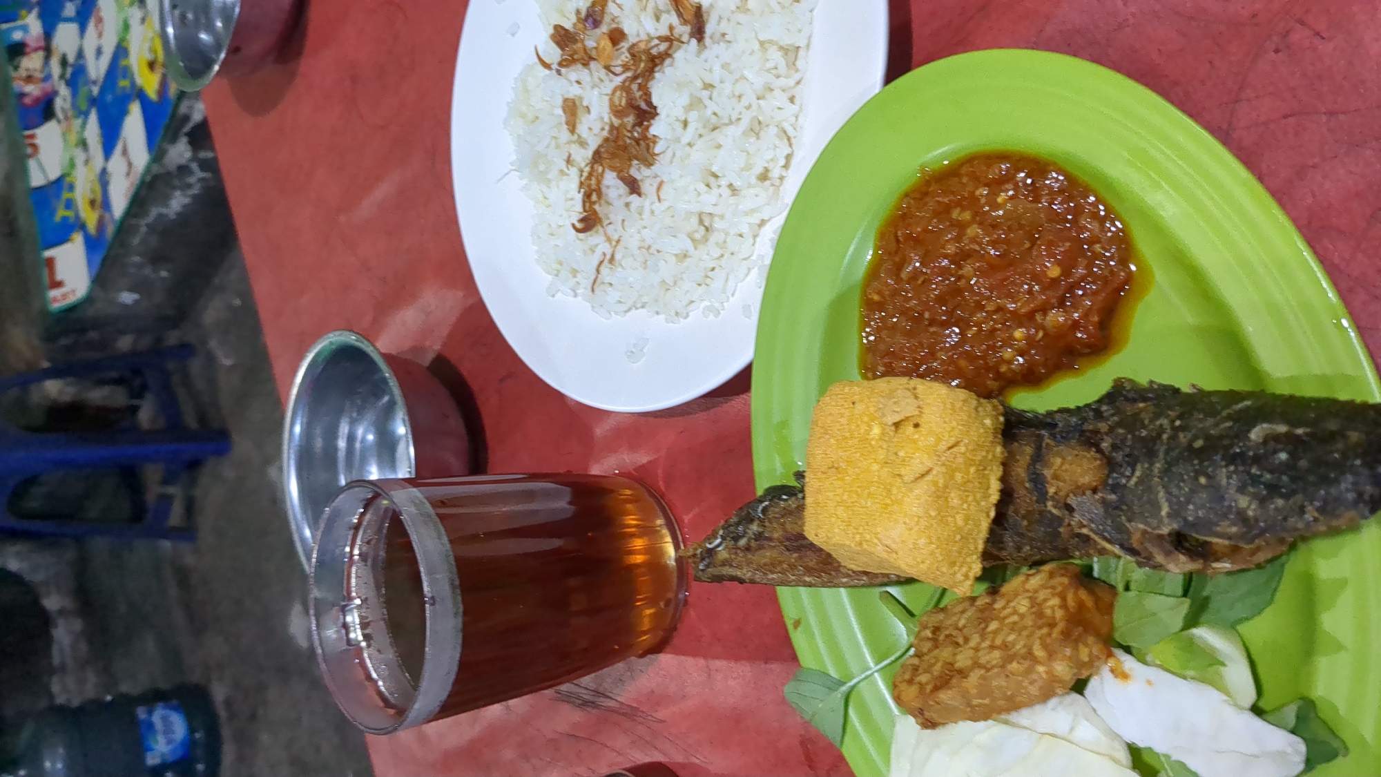 A close-up, eye-level shot captures a meal served on a red table, likely in Purwakarta, Indonesia. In the foreground, a bright green plate holds a portion of fried fish, a golden square of fried tofu, a piece of fried tempeh, a dollop of reddish chili paste, and a few pieces of raw cabbage. To the left, a clear glass is filled with a dark amber liquid, presumably tea. Behind the glass, a small metal bowl sits on the table. In the background, a white plate contains a mound of white rice topped with fried shallots. The overall setting suggests a casual dining environment, possibly an outdoor or simple eatery. The lighting is dim, creating a slightly shadowy atmosphere.