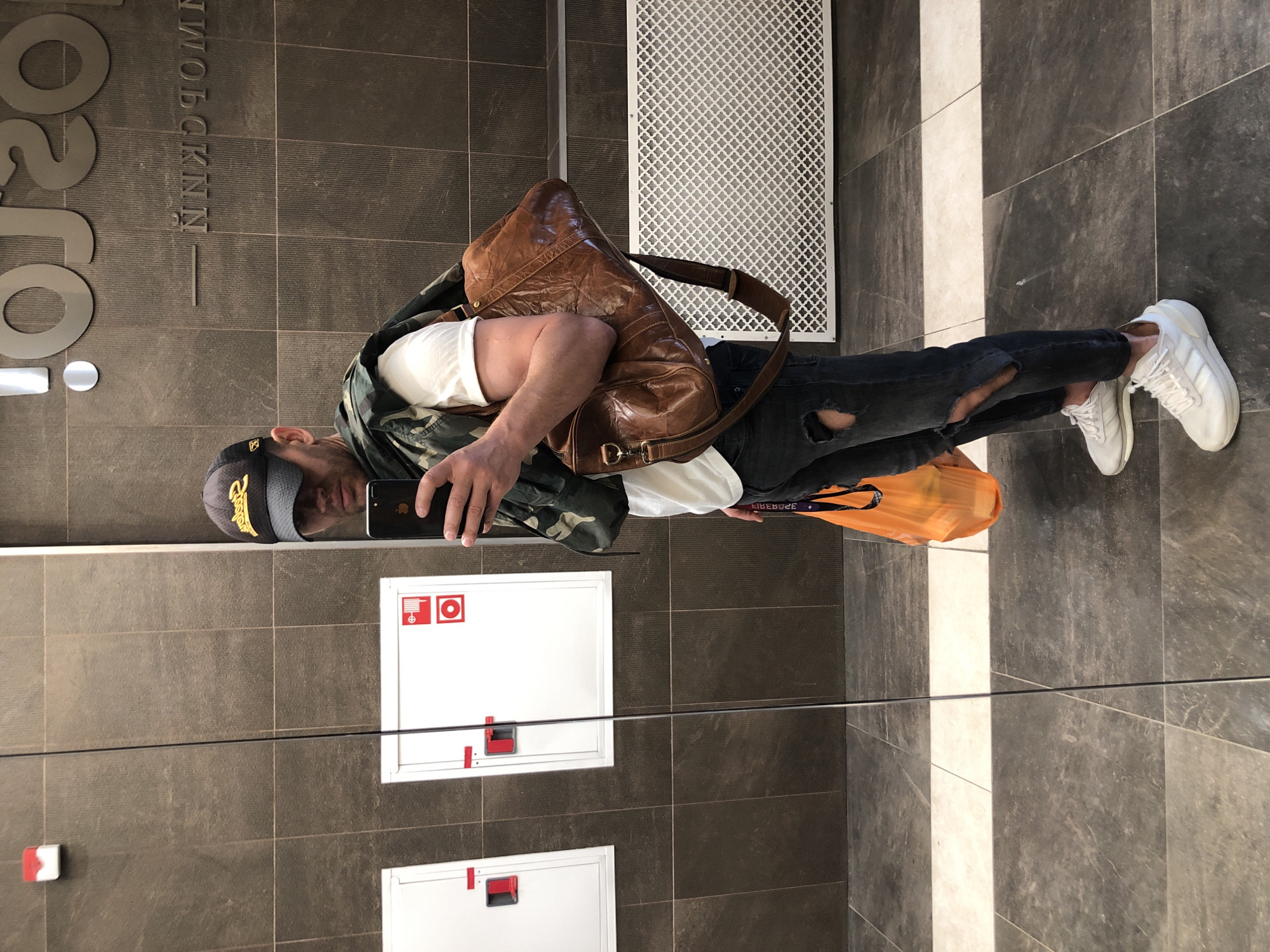 A man stands in a reflective hallway, taking a mirror selfie. He is wearing a black baseball cap with a yellow logo, a white t-shirt, ripped dark jeans, and white sneakers. A camouflage jacket is draped over his shoulders, and he carries a brown leather duffel bag. In his right hand, he holds a smartphone and an orange plastic bag. The hallway features dark tiled walls, white cabinets with red handles on the left, a decorative metal grate on the right, and a tiled floor with a reflective strip. The reflection in the mirror shows a portion of the man's face and the signage "OJO" and "NNKPOMM" on the wall behind him, suggesting a commercial or public space, possibly a mall or a transit hub, in Saint Petersburg, Russia.