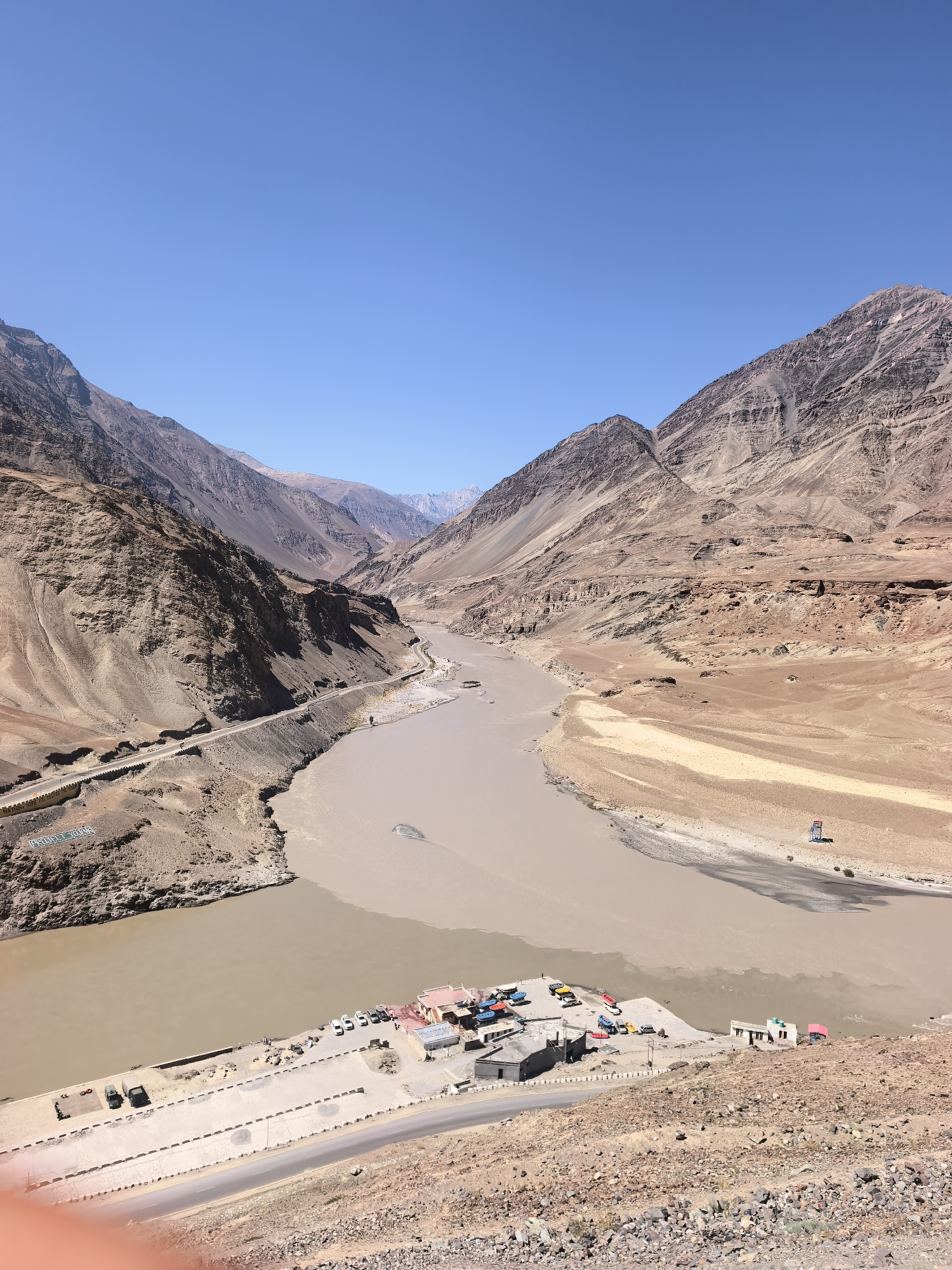 The image depicts a wide, muddy river flowing through a vast, arid canyon under a clear blue sky. The canyon walls are steep and rocky, with shades of brown and tan dominating the landscape. A paved road runs along the left bank of the river, bordered by a low concrete barrier. On the right bank, a small settlement with several buildings and a parking area is visible. Colorful boats are docked near the shore. A few vehicles, including cars and what appear to be military trucks, are parked in the settlement's lot. The river itself is a prominent feature, with a distinct change in color where two tributaries meet, suggesting different sediment loads. The overall scene is one of natural grandeur and remoteness.