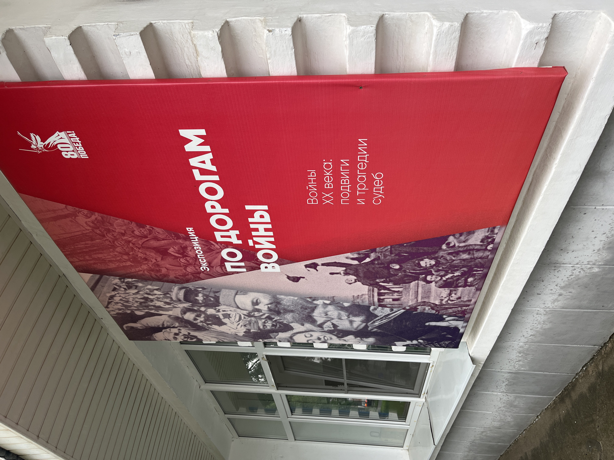A large, red banner with white text and graphics is mounted on the exterior wall of a building. The banner advertises an exhibition titled "By the Roads of War." On the upper right portion of the banner, there is a white graphic of a winged female figure raising a sword, with the number "80" and the text "VICTORY!" below it. The banner also features black and white historical photographs on the left side. Text on the banner reads "EXHIBITION," "BY THE ROADS OF WAR," and "Wars of the 20th Century: Feats and Tragedies of Fates." The banner is partially obscuring a row of windows. The building appears to be white and has a textured, layered facade. In the background, through the windows, trees and what appears to be another building can be seen. The banner is displayed on a building in the city of Chaikovsky, Russia, likely as part of a public event or commemoration.