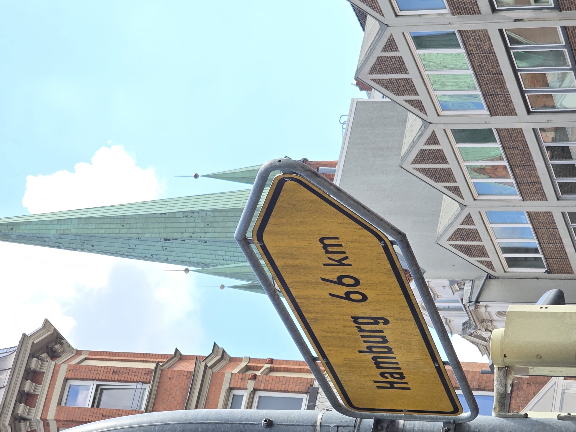 An eye-level, medium shot captures a yellow directional signpost in Lübeck, Germany, pointing towards "Hamburg 66 km." The signpost is mounted on a metal pole and features a black border. Behind the sign, a tall, green, spire-like structure, likely a church steeple, dominates the background. To the left of the steeple, a red brick building with white-trimmed windows is visible. In the foreground, a light-colored, cylindrical object is mounted on the same pole as the signpost. The sky is bright blue with scattered white clouds. The overall scene depicts an urban environment, likely on a sunny day.