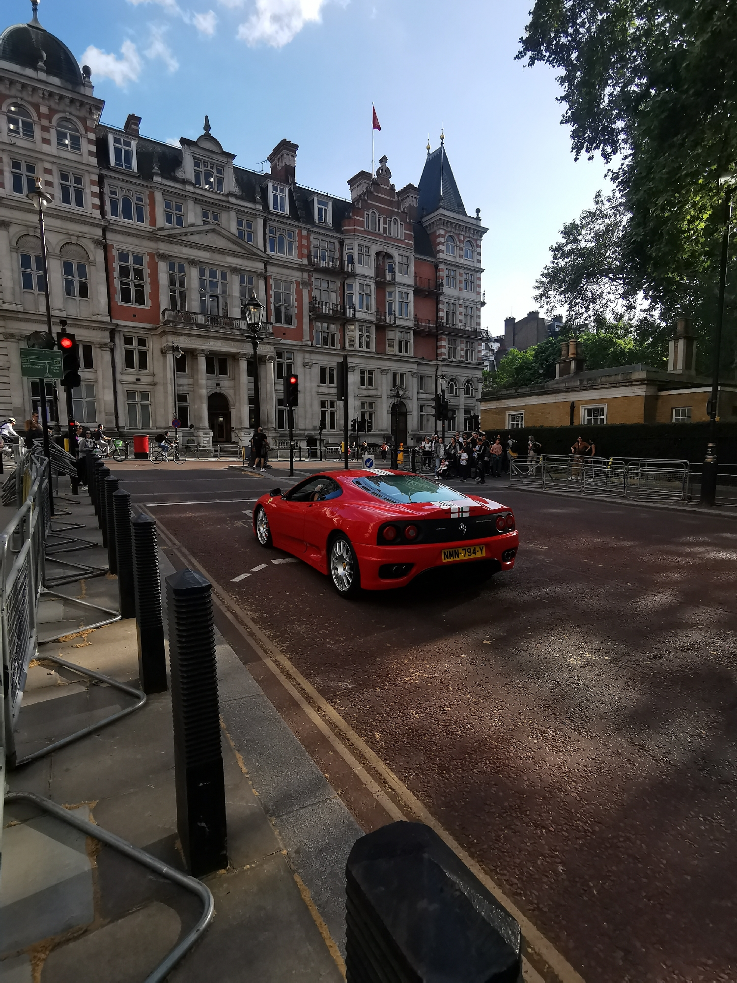 This daytime street scene captures a bustling intersection in London, United Kingdom, under a bright blue sky with scattered white clouds. Strong sunlight casts distinct shadows, suggesting late morning or early afternoon.
The central subject is a striking red Ferrari, likely a 360 Modena, viewed from the rear three-quarter angle. It features a white racing stripe across its engine cover and a visible UK license plate "NMN 794 Y". The car is positioned on an asphalt road, which has sections of reddish-brown pavement, potentially indicating a bus or cycle lane. Double yellow lines run along the curb, and white dashed lines mark the road.
On the left, a grand, multi-story building dominates the background. This is the Institution of Civil Engineers (One Great George Street), identified by the partially visible text "INSTITUTE OF CIVIL ENGINEERS" above its main entrance. The building showcases ornate classical architecture with red brickwork, light stone facades, numerous windows, balconies, intricate roof details, turrets, and a small red flag on its pinnacle.
Several people are present on the sidewalks: some are walking, others are standing behind silver crowd control barriers and black bollards. A cyclist is also visible. Two red traffic lights are illuminated, indicating traffic control at the intersection. To the right, lush green trees suggest a nearby park or green space, and behind a hedge, a smaller, light-colored building can be seen. Further affirming the London location are a green street sign partially visible on the left, indicating "Westminster / Hyde Park," and a classic red telephone box in the background.