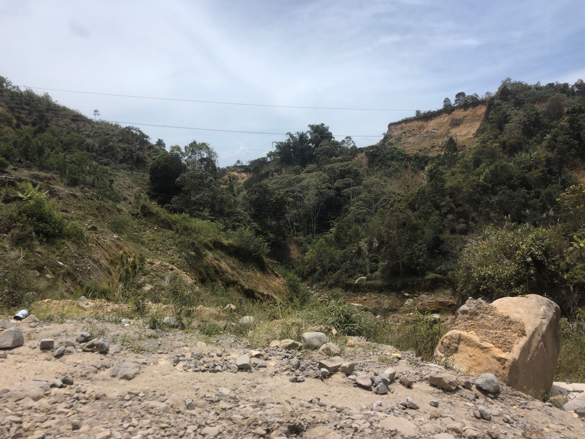The image depicts a rocky, uneven terrain with dense vegetation covering the surrounding hills. A prominent landslide scar is visible on the right side of the frame, revealing layers of soil and rock. Several power lines stretch across the sky, indicating proximity to infrastructure. The foreground is dominated by gravel and scattered rocks, with a large, broken boulder on the right. In the mid-ground, various types of foliage and trees populate the slopes, with evidence of erosion and exposed earth. A few plastic bottles and a white plastic bag are discarded on the ground, suggesting human activity in the area. The sky is partly cloudy, casting a diffused light over the scene. This landscape is indicative of the natural environment found in Bintangpermata, Indonesia, a region known for its mountainous terrain and susceptibility to landslides.