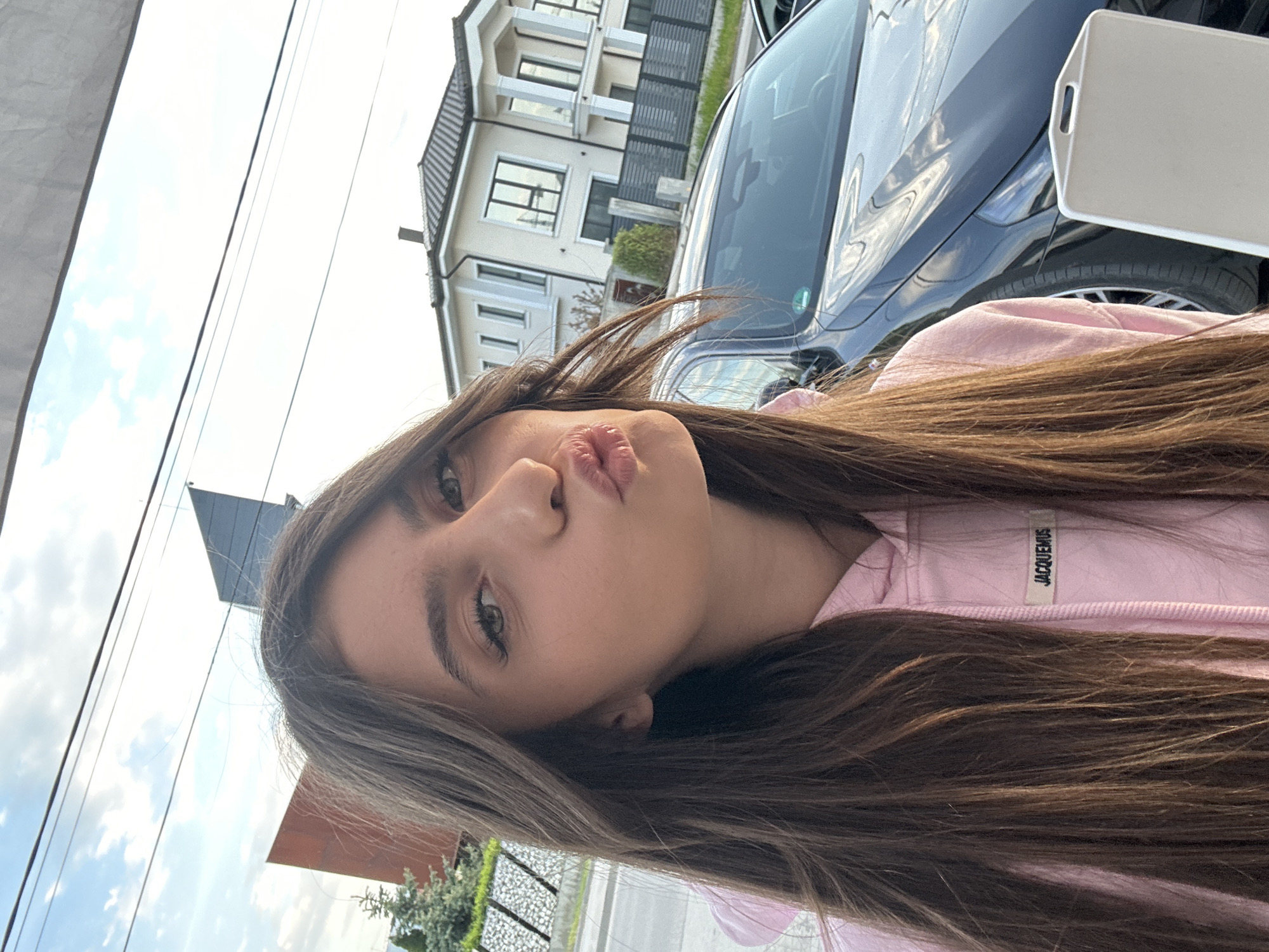 The image features a young woman posing for a close-up photo, with her head tilted to the side and her lips pursed in a "kiss" face. Her long, brown hair falls over her shoulders and chest. She is wearing a pink top, and a white tag with the brand name "JACQUEMUS" is visible on the neckline.
In the background, a dark blue or black sedan is partially visible to the right of the woman. Behind the car and extending across the upper left, there is a modern, multi-story house with light-colored exterior walls and dark-tiled roofs. Various windows and balconies are discernible on the building. Above the house and car, the sky is bright with scattered white clouds. Several thin, dark utility lines crisscross the sky. Some green trees or bushes are also visible near the base of the house and in the lower left portion of the image.
The photo appears to be taken outdoors during daytime, indicated by the natural lighting and clear sky. The setting suggests a residential area, consistent with the provided location context of Plovdiv, Bulgaria. The overall image is rotated 90 degrees counter-clockwise.
