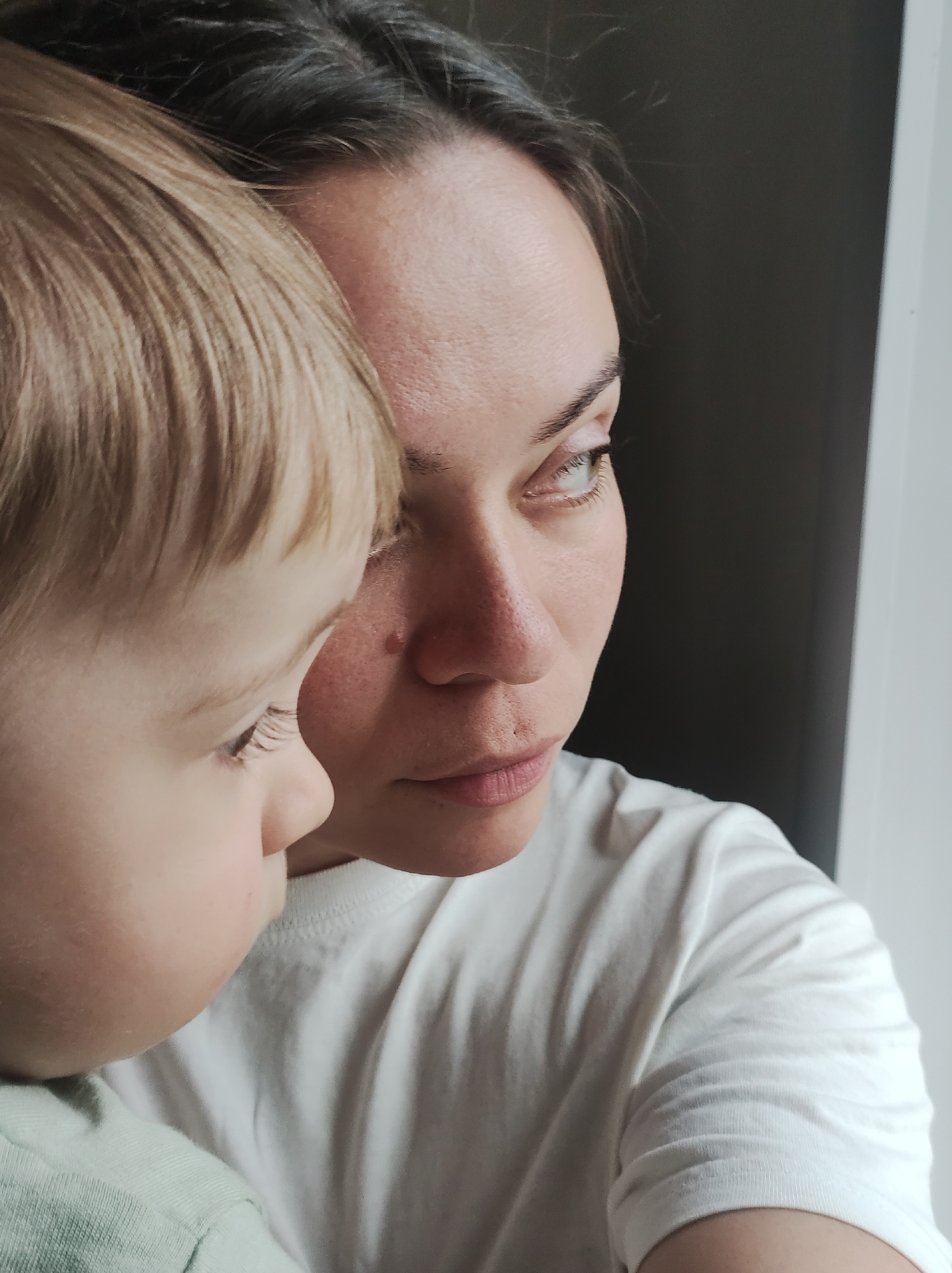 A close-up shot features a fair-skinned woman and a young child, potentially a toddler, positioned close together. The woman has dark hair, light eyes, and is wearing a white t-shirt. She is gazing slightly upward and to the right, with a thoughtful expression. The child, who has light blond hair, is positioned on the left side of the frame, with the side of their face visible. They seem to be looking down. Both figures are positioned near a window frame visible on the right side of the frame. The location is Unknown, Unknown.