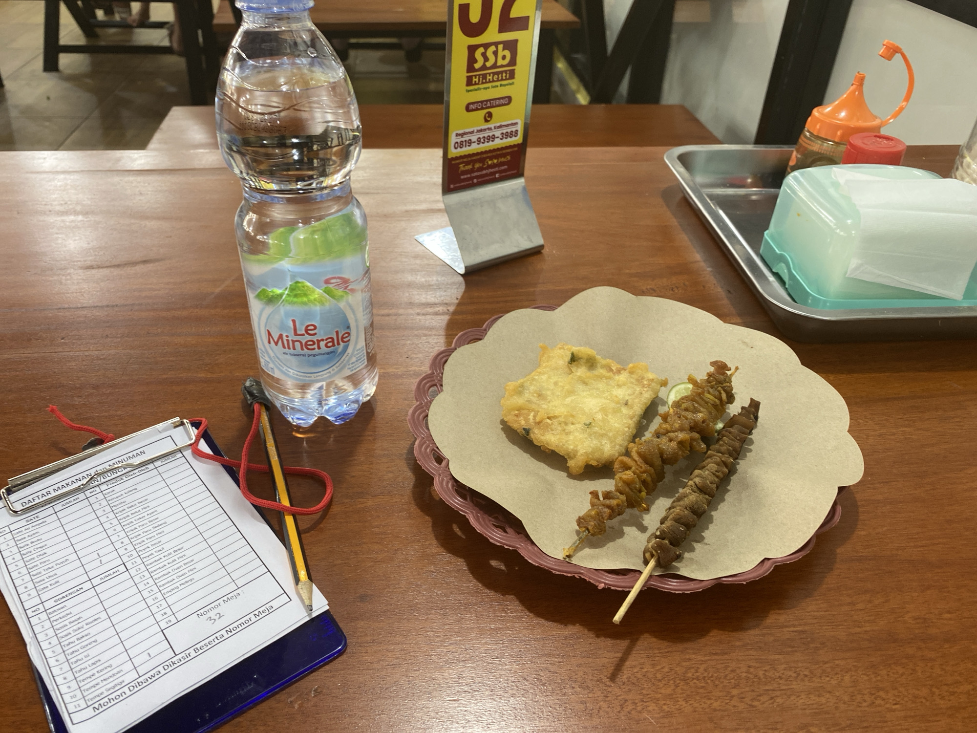 The image depicts a close-up overhead view of a wooden table in what appears to be an outdoor dining setting, likely at a restaurant, in North Jakarta, Indonesia. A bottle of "Le Minerale" mineral water stands on the left side of the table, next to a clipboard with a pen and a pencil. In the center, a plate holds a piece of fried food, possibly a fritter, and two skewers of grilled meat or seafood. To the right, a metal tray contains a dispenser of tissues, a red-lidded container, and an orange squeeze bottle, likely for condiments. A stand with a yellow and red sign, advertising "SSb Hj. Hesti" and providing contact information for "INFO CATERING," is visible in the background. The overall scene suggests a casual meal being prepared or consumed, with the ordering process documented on the clipboard.